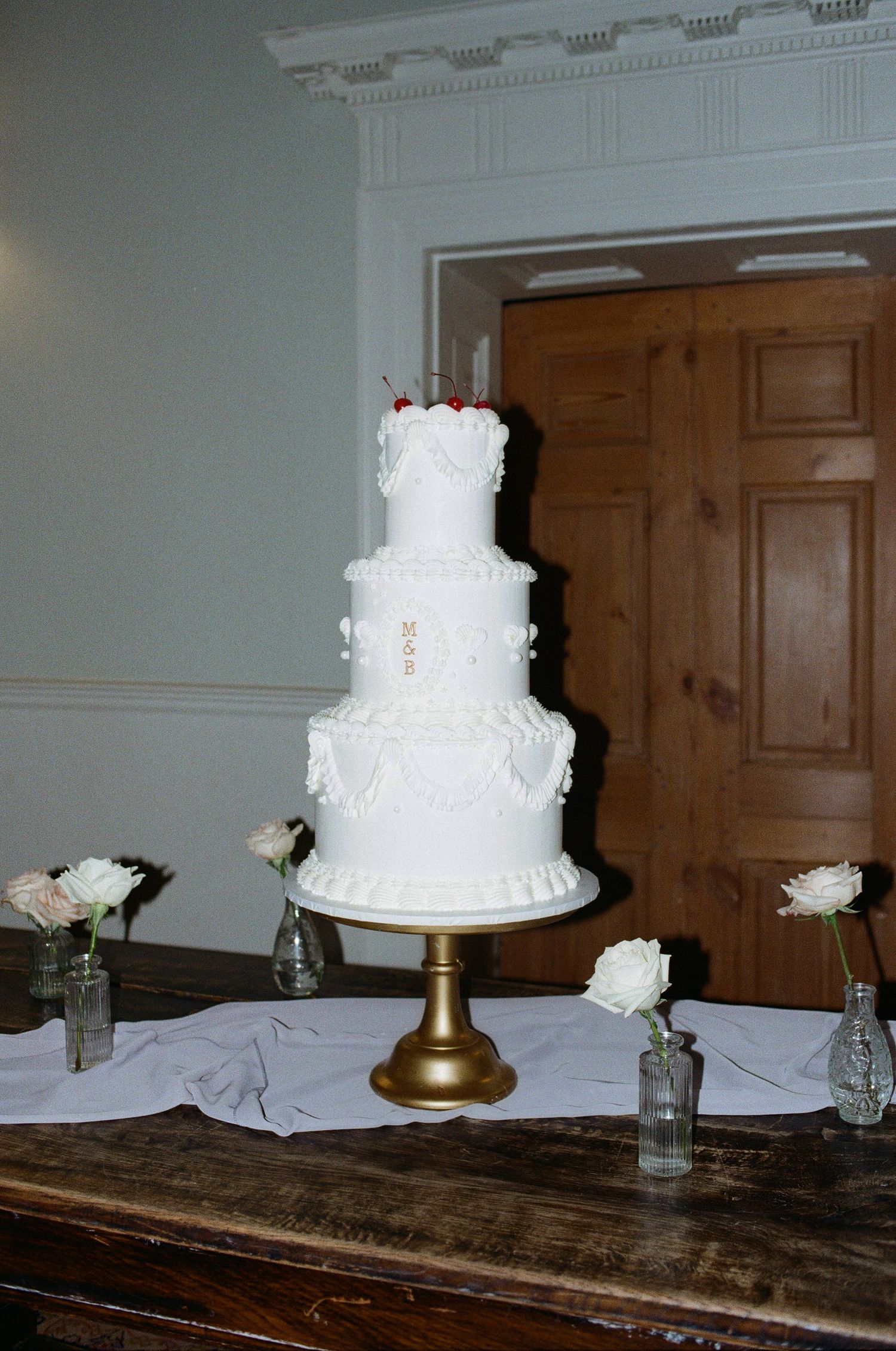 A three-tiered white wedding cake decorated with lace details sits on a gold stand with white roses in glass vases as decor.