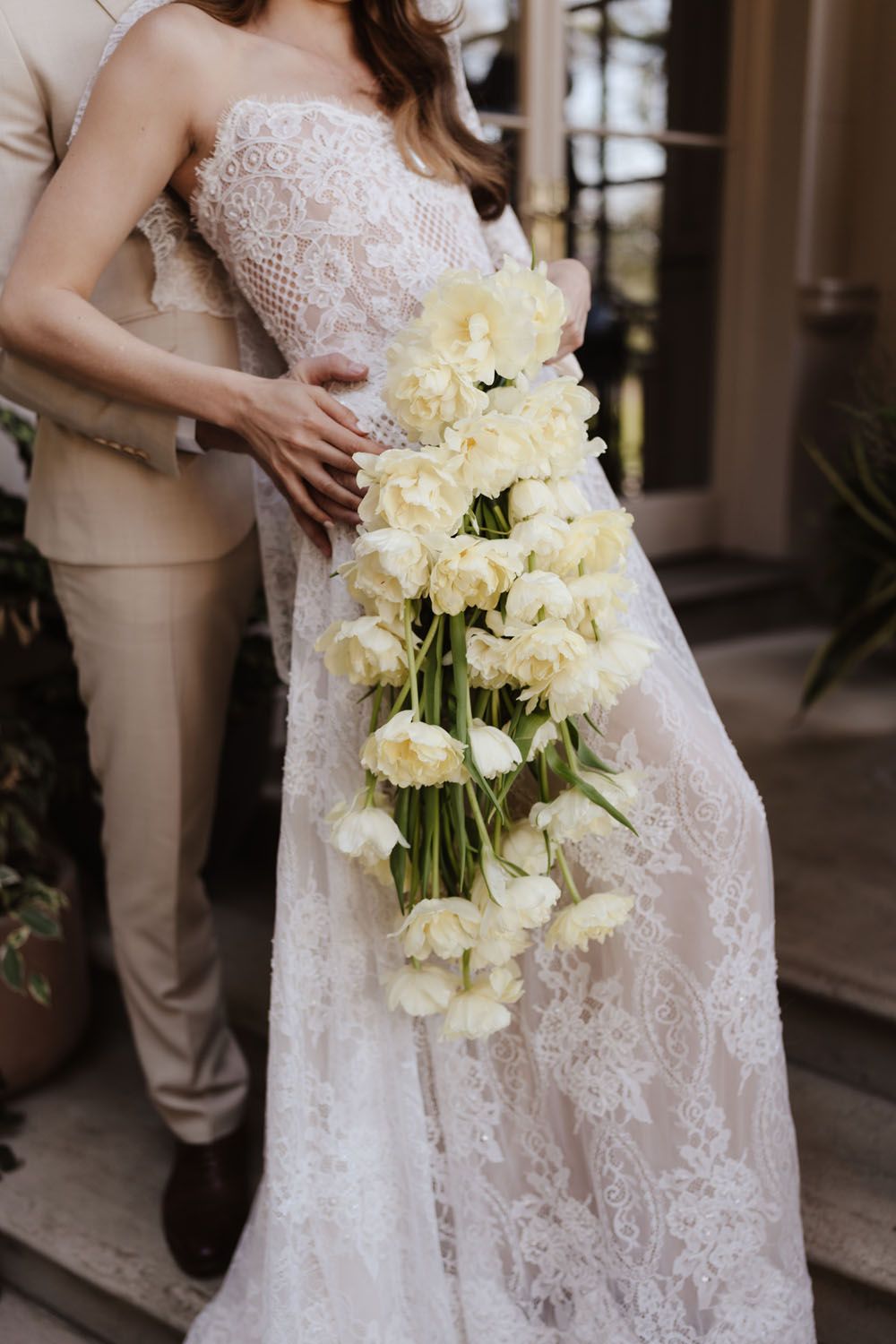 Bride in lace gown holding cascading white tulip bouquet with groom in beige suit at wedding ceremony.