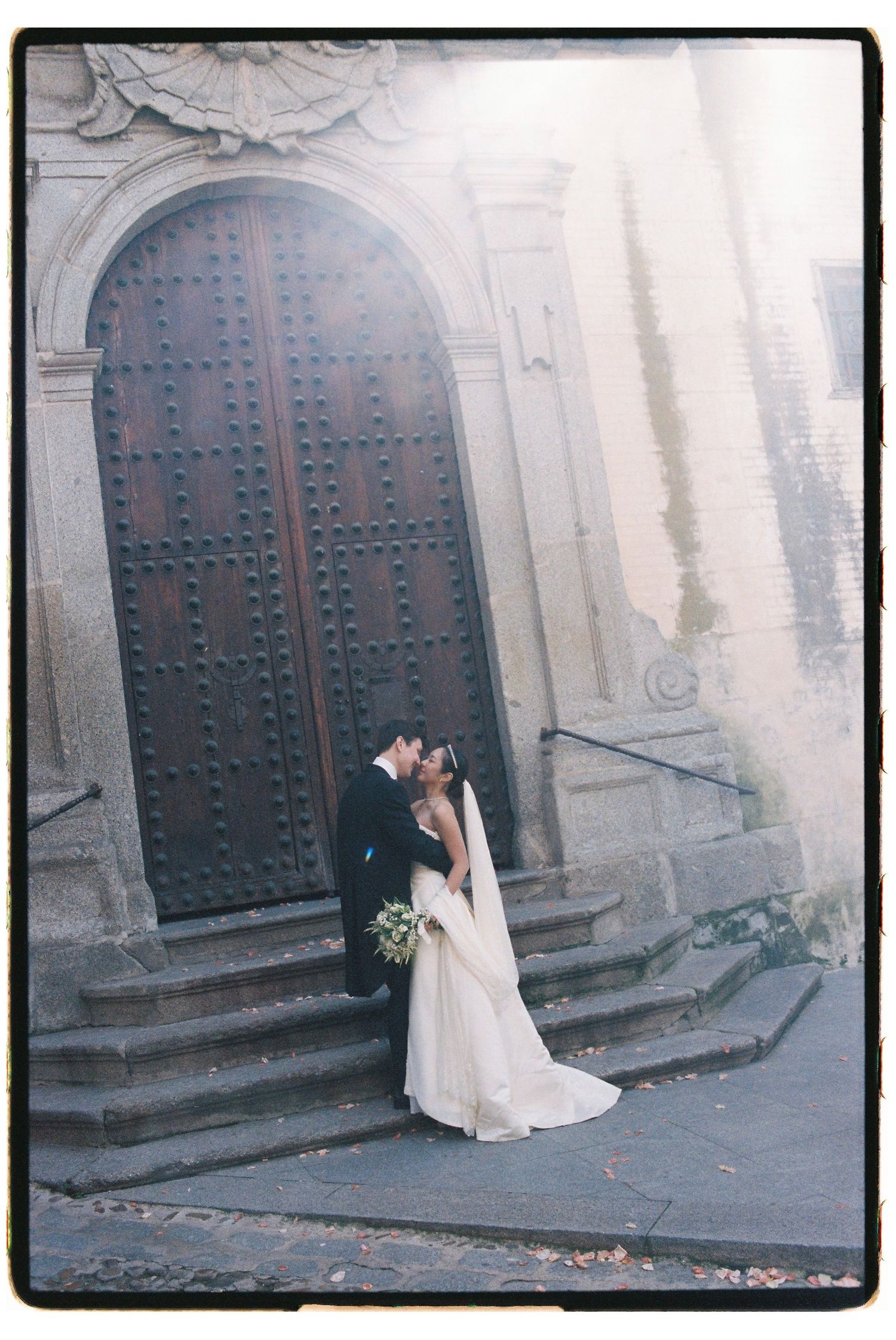 Bride and groom sharing a kiss before a grand arched wooden door with stone steps.