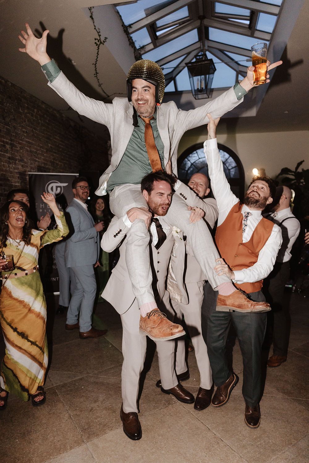 Wedding celebration at an indoor venue with people dancing and one person being lifted up during a festive moment.