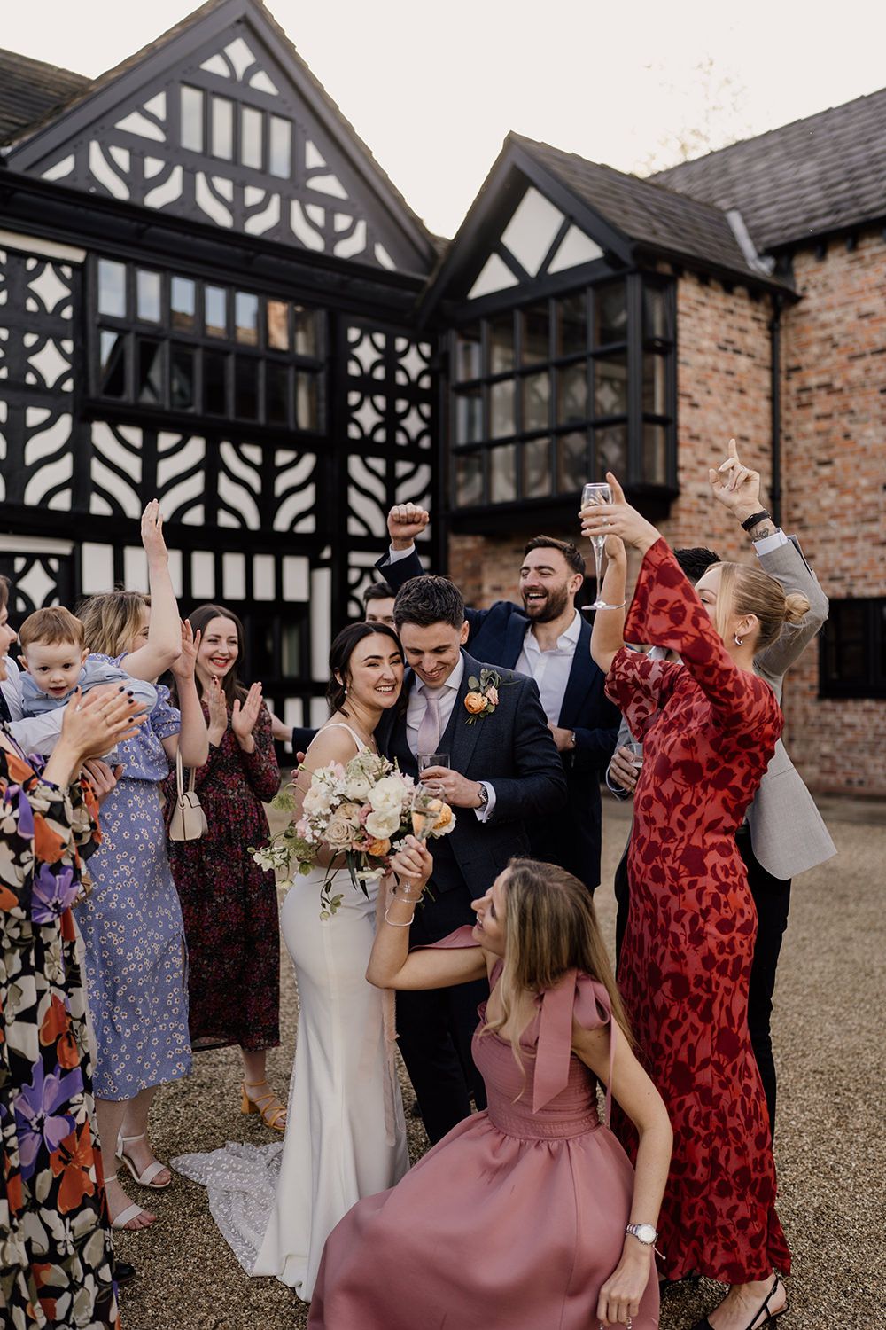 Bride and groom kissing as friends dressed in bright wedding outfits laugh and dance with Holford Hall behind them.