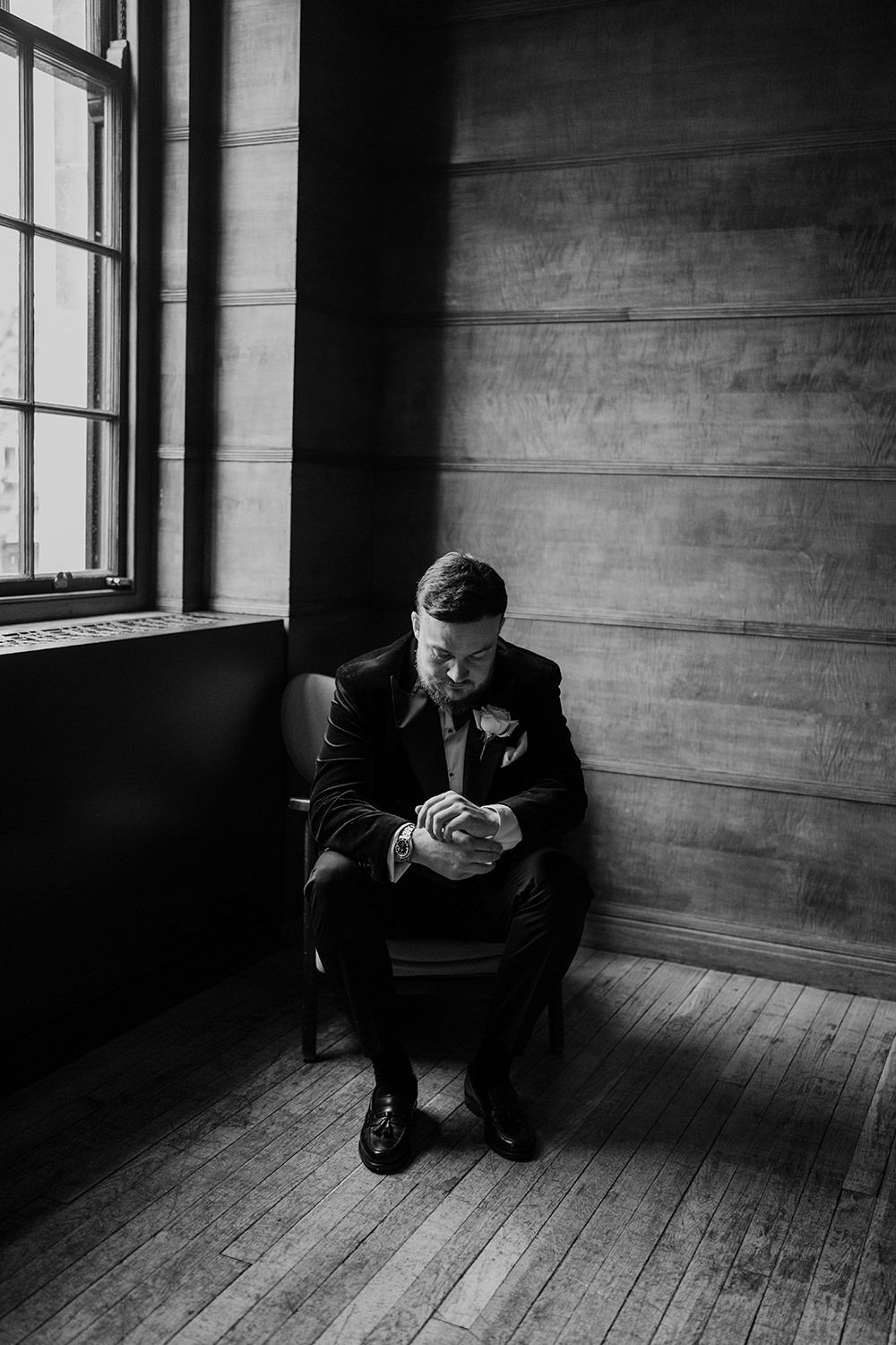 Black and white photo of a person in a suit sitting alone in an empty room by a window looking down.