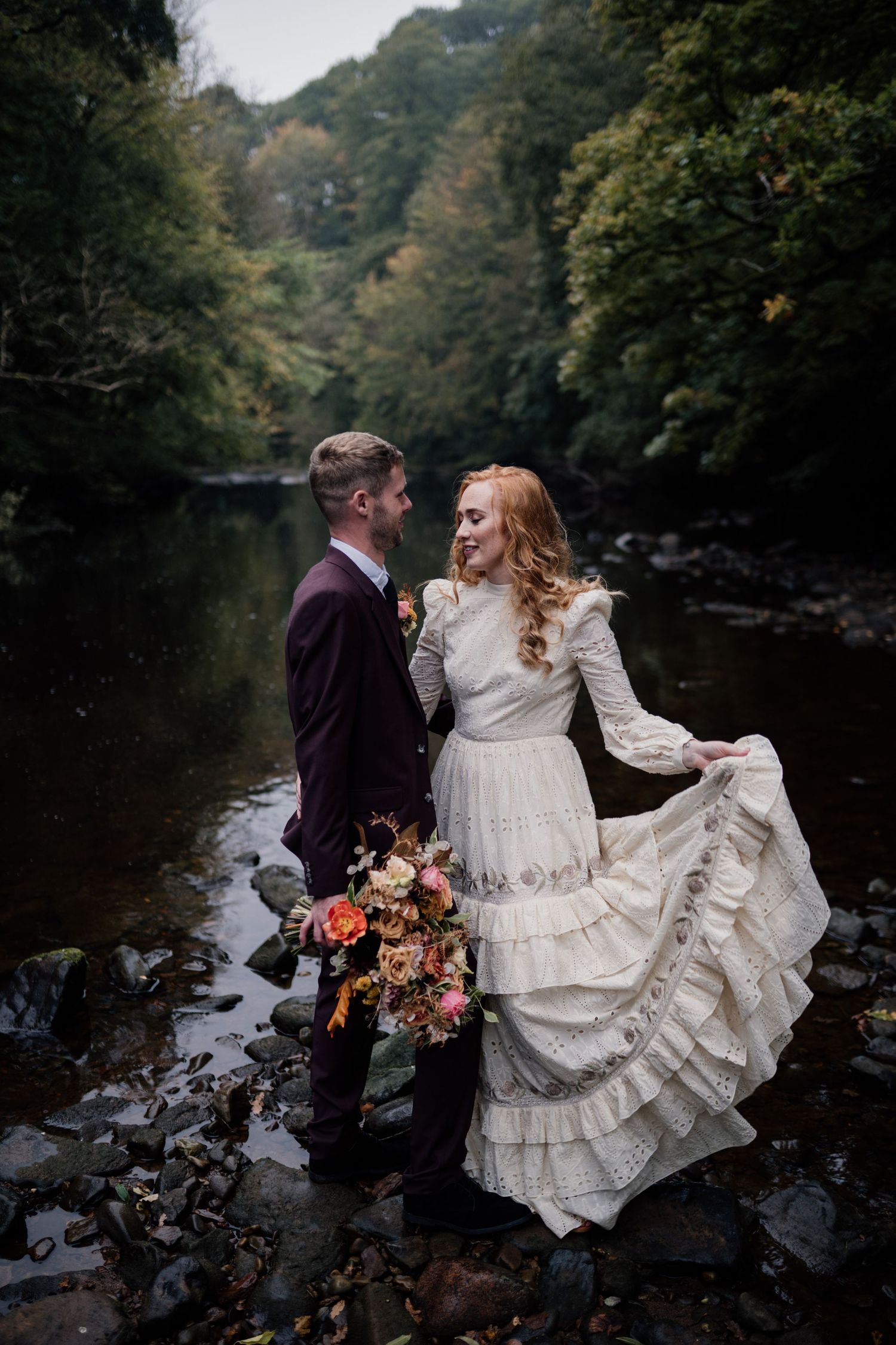 Couple smiling on their wedding day, stood on rocks on a river with trees surrounding them.