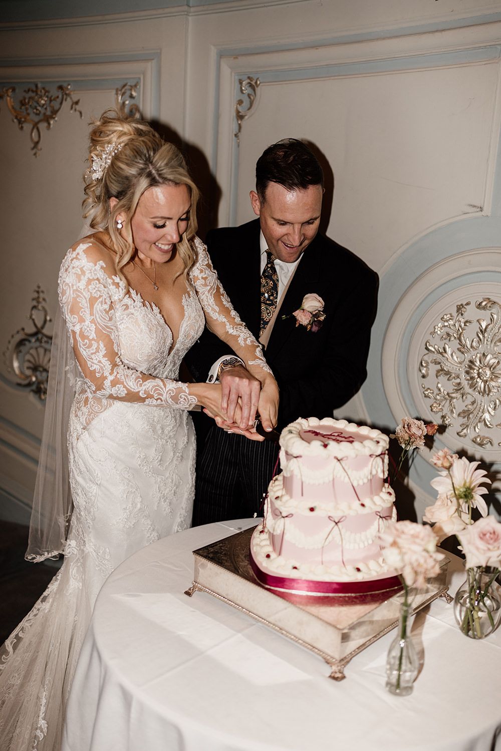 A newlywed couple cuts their wedding cake together while smiling in an ornately decorated room.