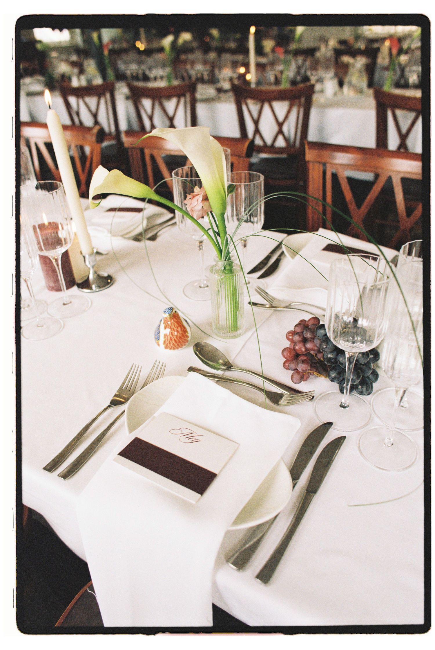 Elegant wedding reception table set with white linens, calla lily centerpiece, candles, grapes, and formal place settings.
