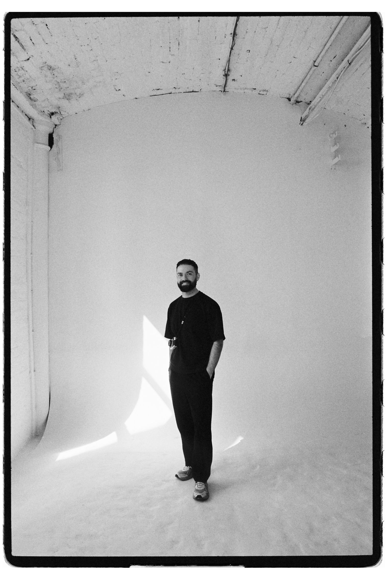 Bearded man standing in bright white studio space in black and white photo.