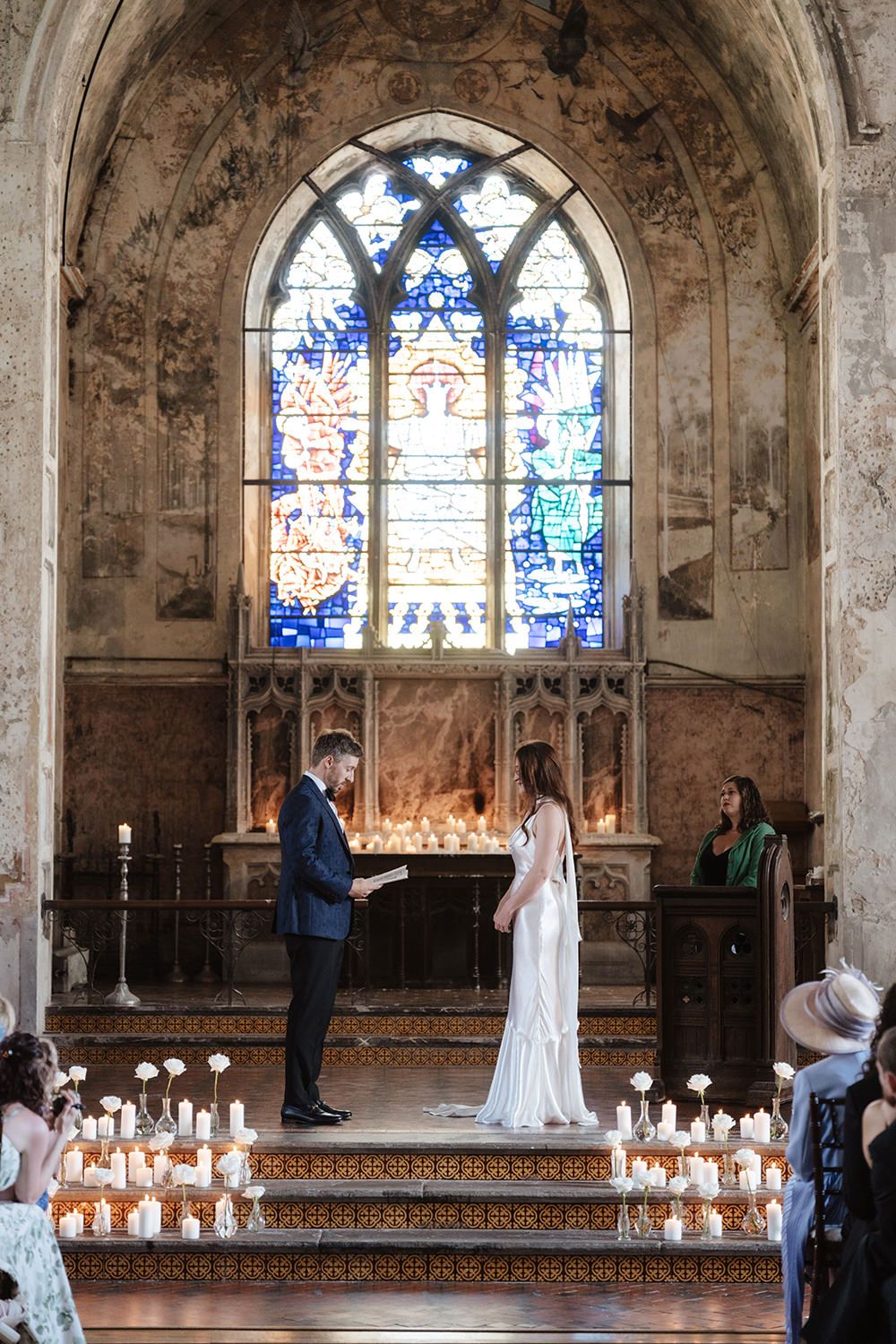 Wedding ceremony in a historic chapel with stained glass windows and rows of white candles lining the aisle.