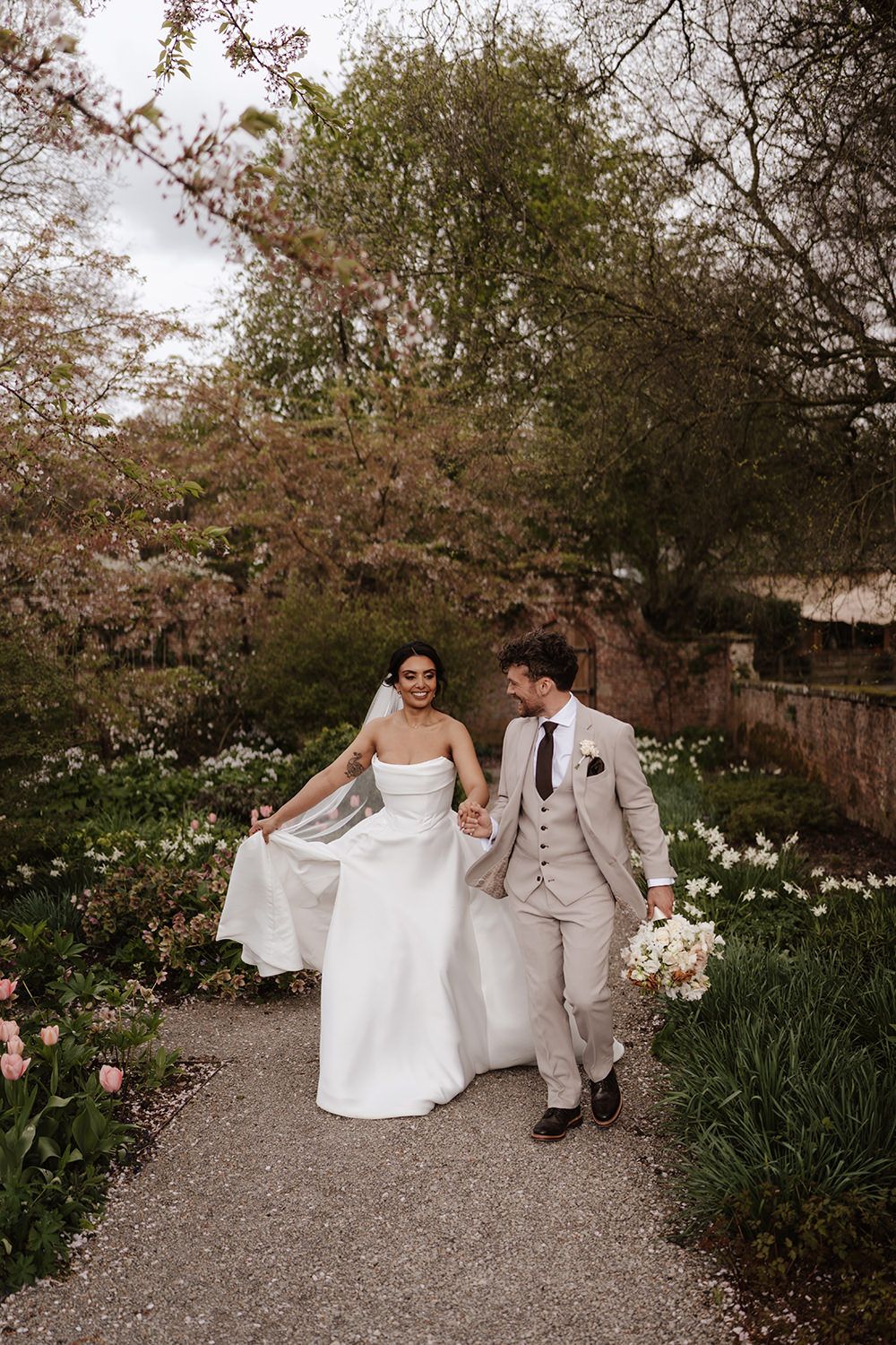 A couple walks along a garden path on their wedding day, surrounded by blooming spring flowers and trees.