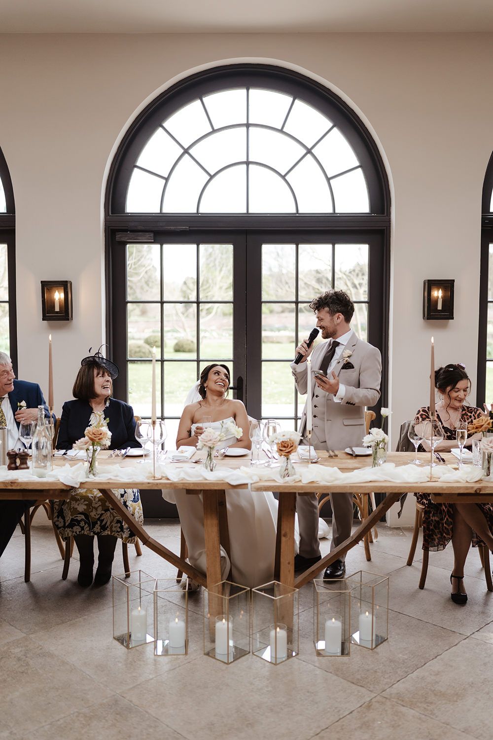 Wedding reception speech at elegant venue with arched window and floral table decor in warm natural lighting.