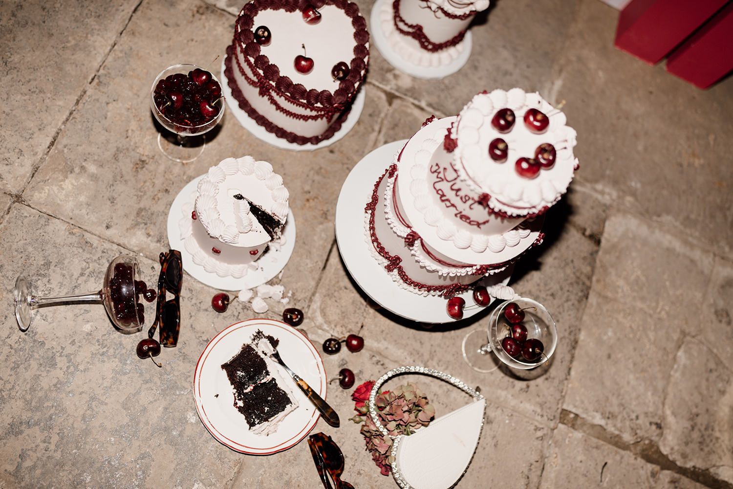 Elegant white wedding cakes decorated with cherries and burgundy accents on a stone floor.