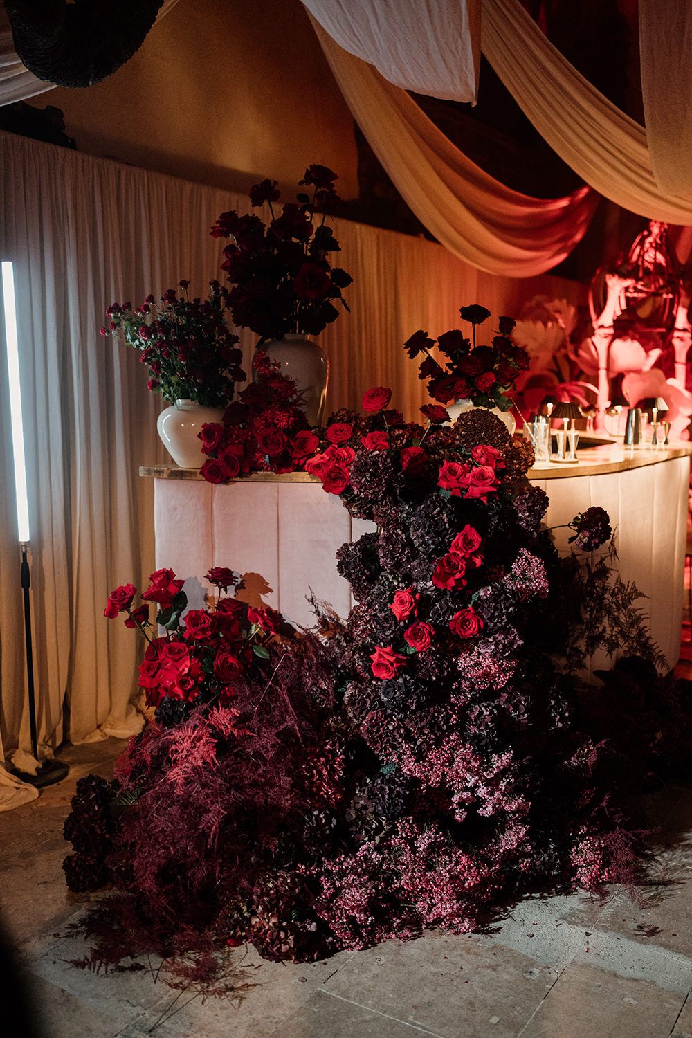 Dramatic floral installation with cascading dark red roses and burgundy foliage against draped white fabric backdrop.