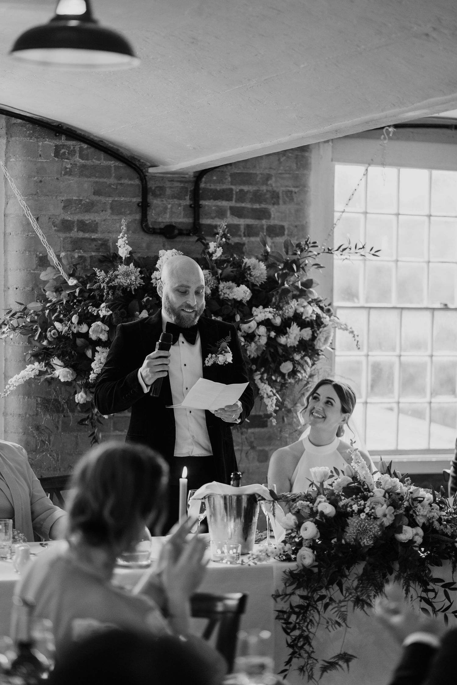 Person giving wedding speech at reception with dramatic lighting and floral backdrop.