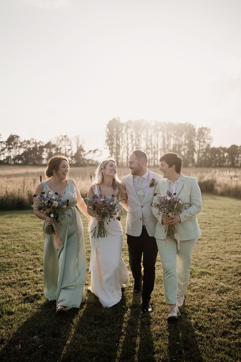 Bride and her bridal party smiling and laughing while walking on grass with the forrest and sunset behind.