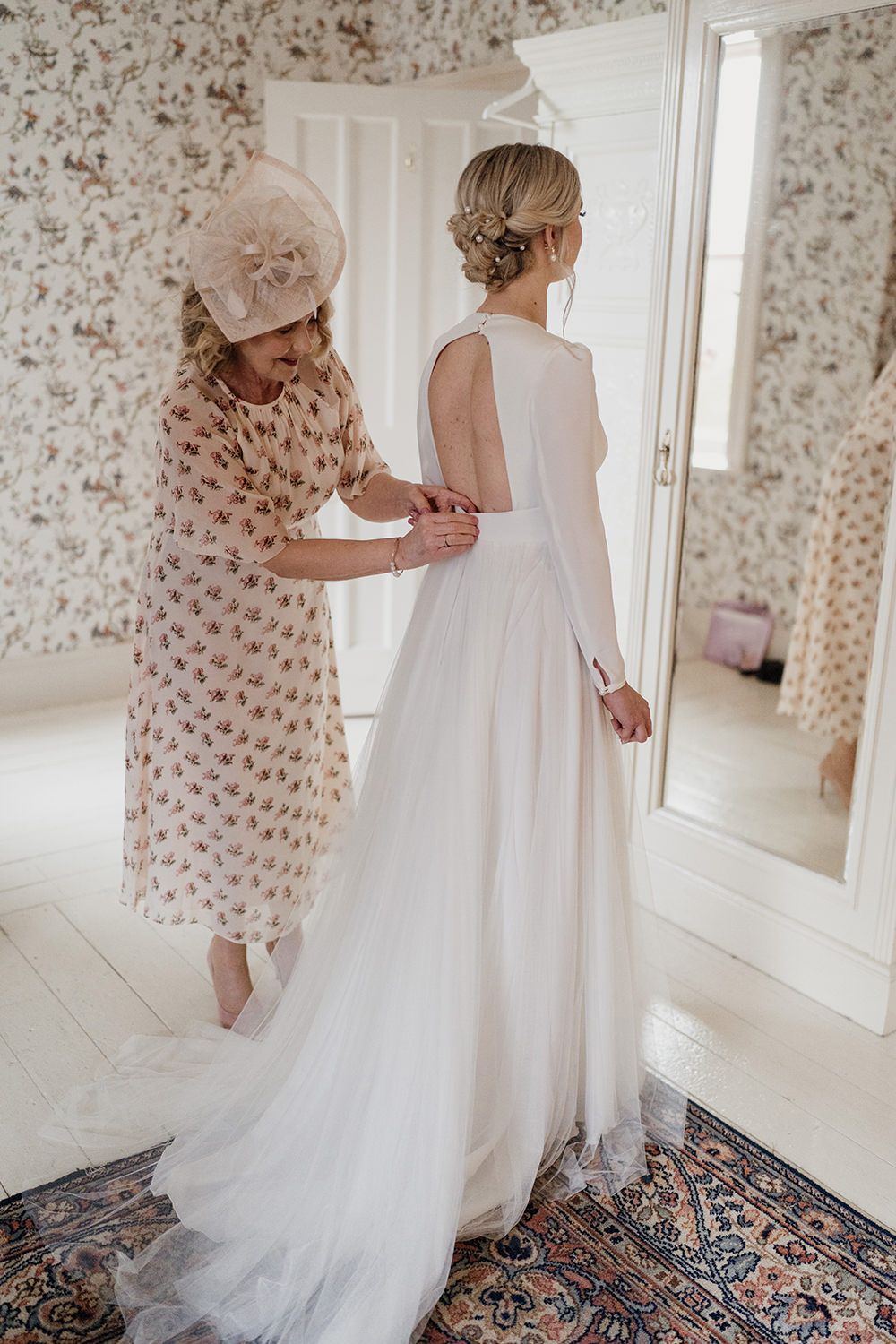 Bride stood as her mother helps to fasten the back of her wedding dress.