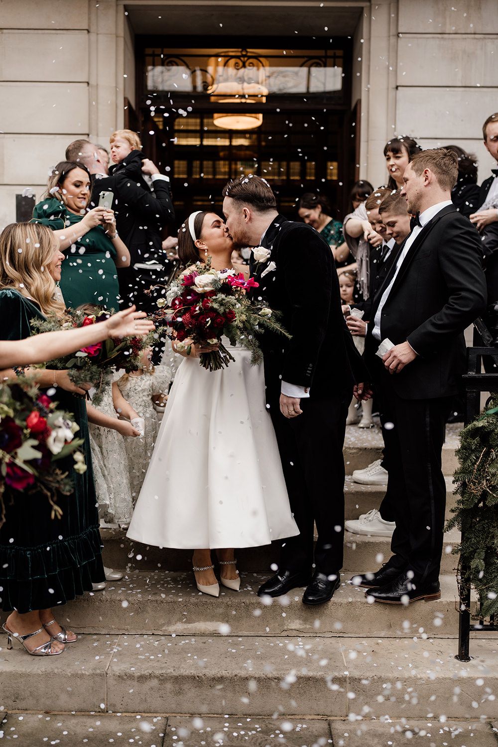 Wedding couple shares a kiss on church steps while guests shower them with confetti during winter celebration.