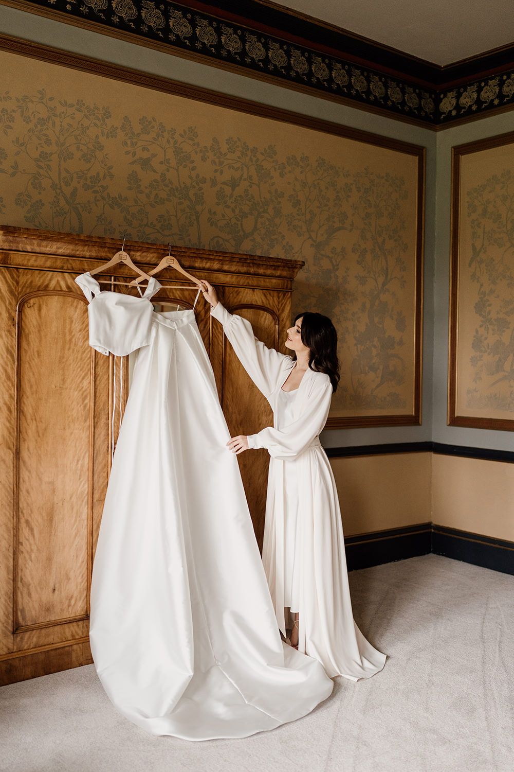 Bride looking and adjusting her wedding dresses hung on a victorian wardrobe.