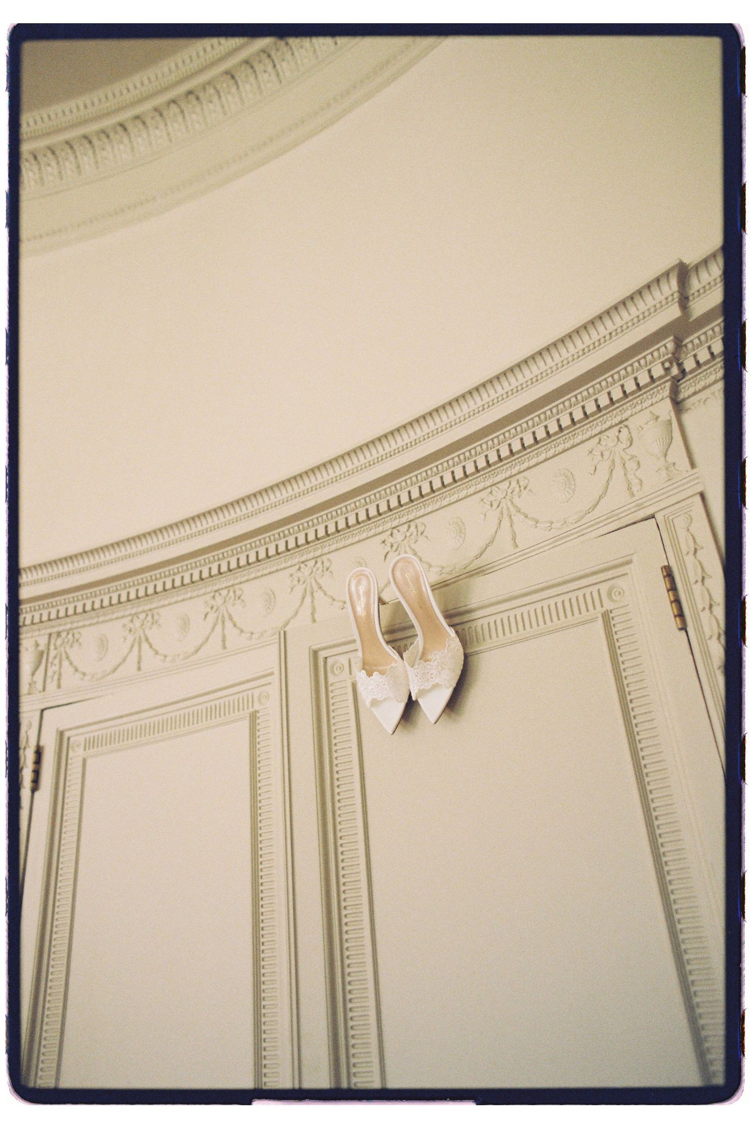 White bridal heels hang delicately on ornate white wall molding in an elegant venue.