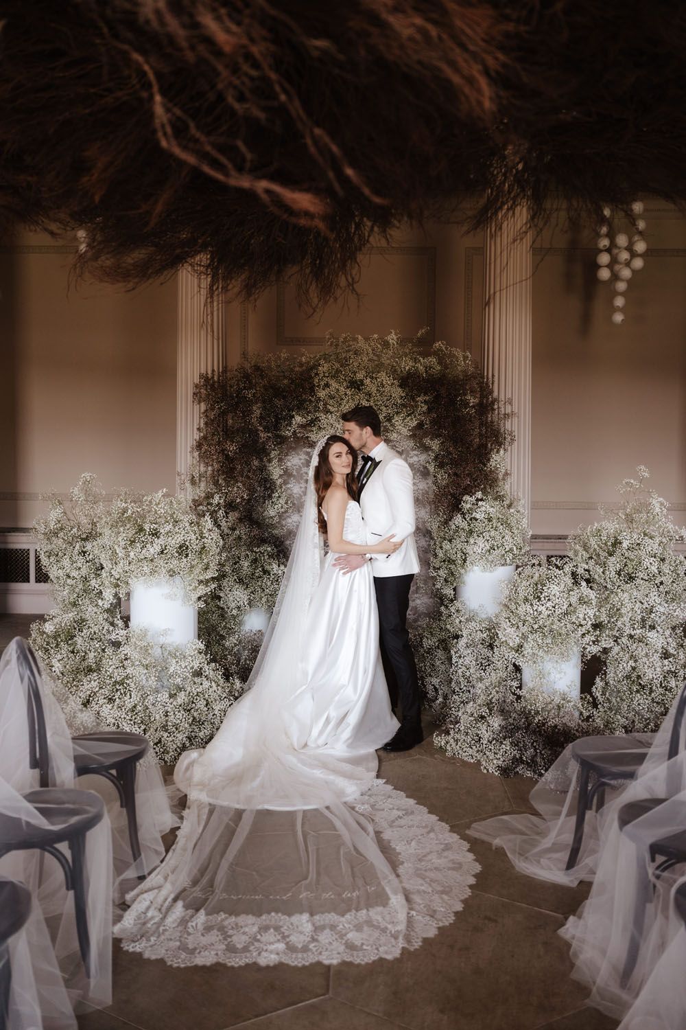 Bride and groom embrace at elegant ceremony altar surrounded by lush greenery and baby's breath floral arrangements.
