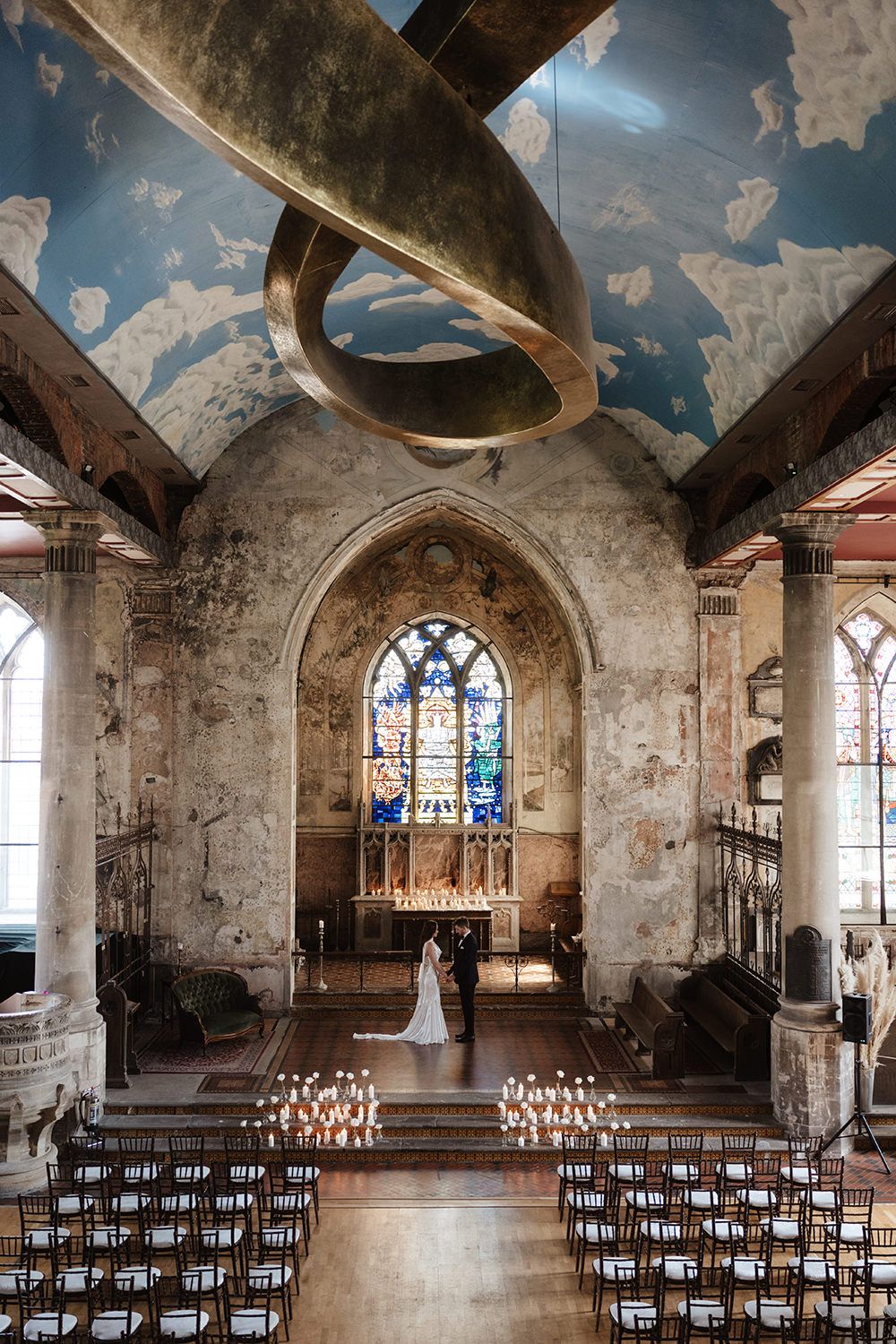 Couple exchanges vows in a candlelit historic church with stained glass windows and a sculptural gold installation overhead.