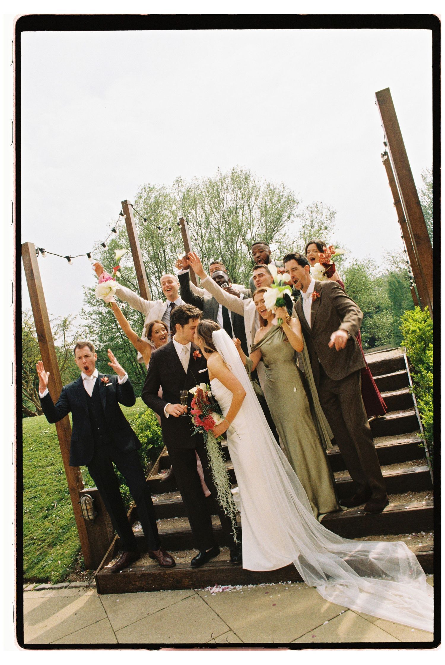 Wedding party celebrating on outdoor steps, bride in white gown with veil, groomsmen and bridesmaids cheering.