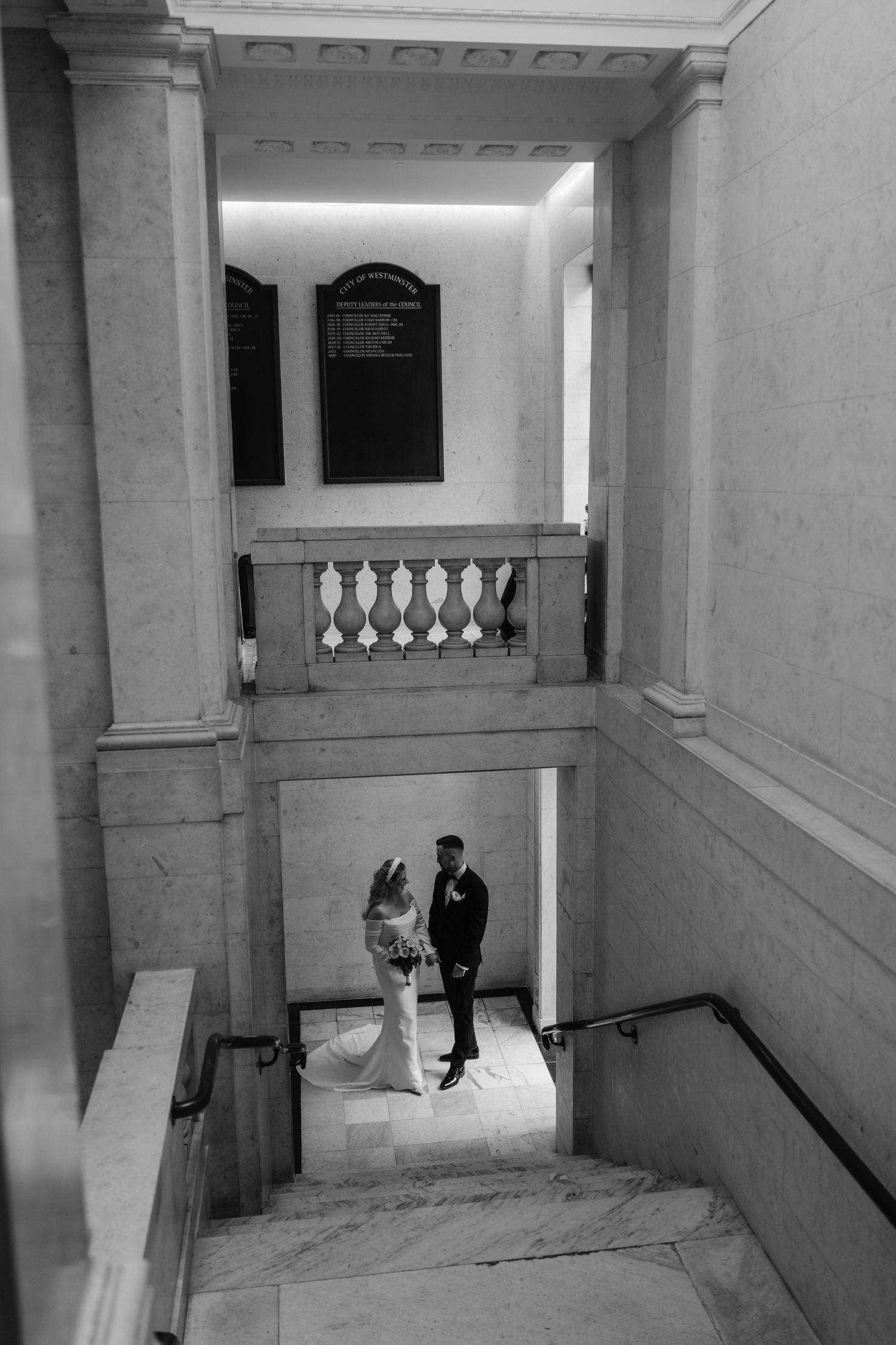 Black and white photo of bride and groom standing on marble staircase in elegant building.