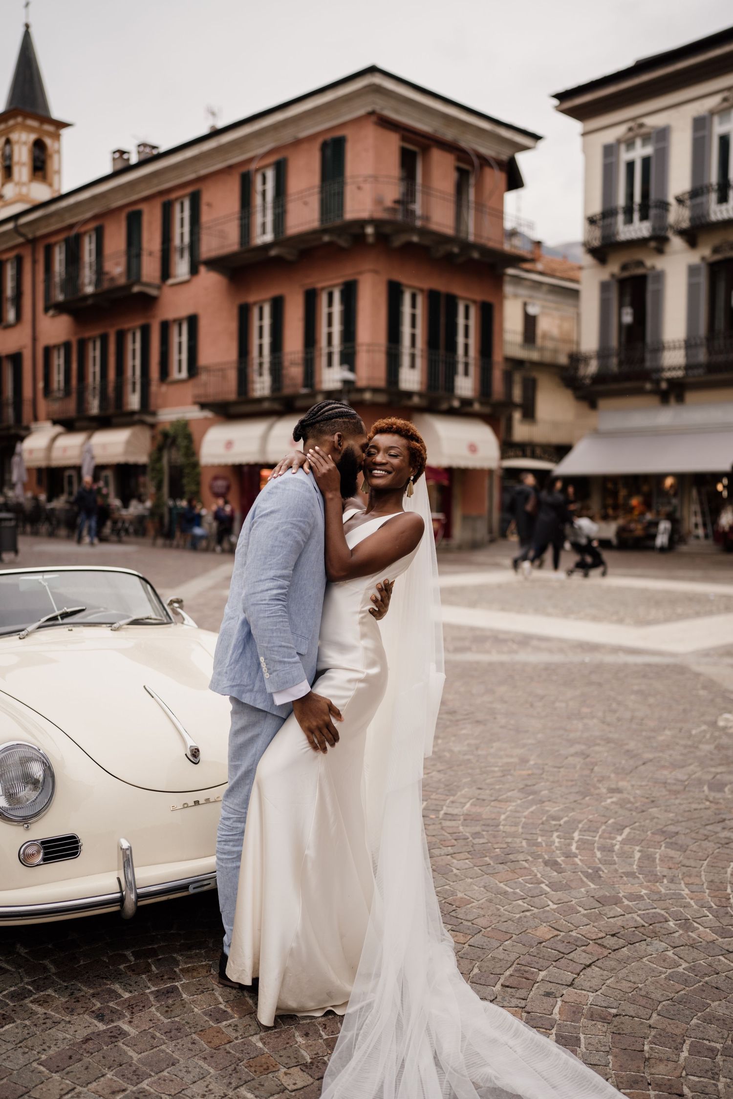 Newlyweds embracing beside a vintage white Porsche in a charming Italian piazza on their wedding day.