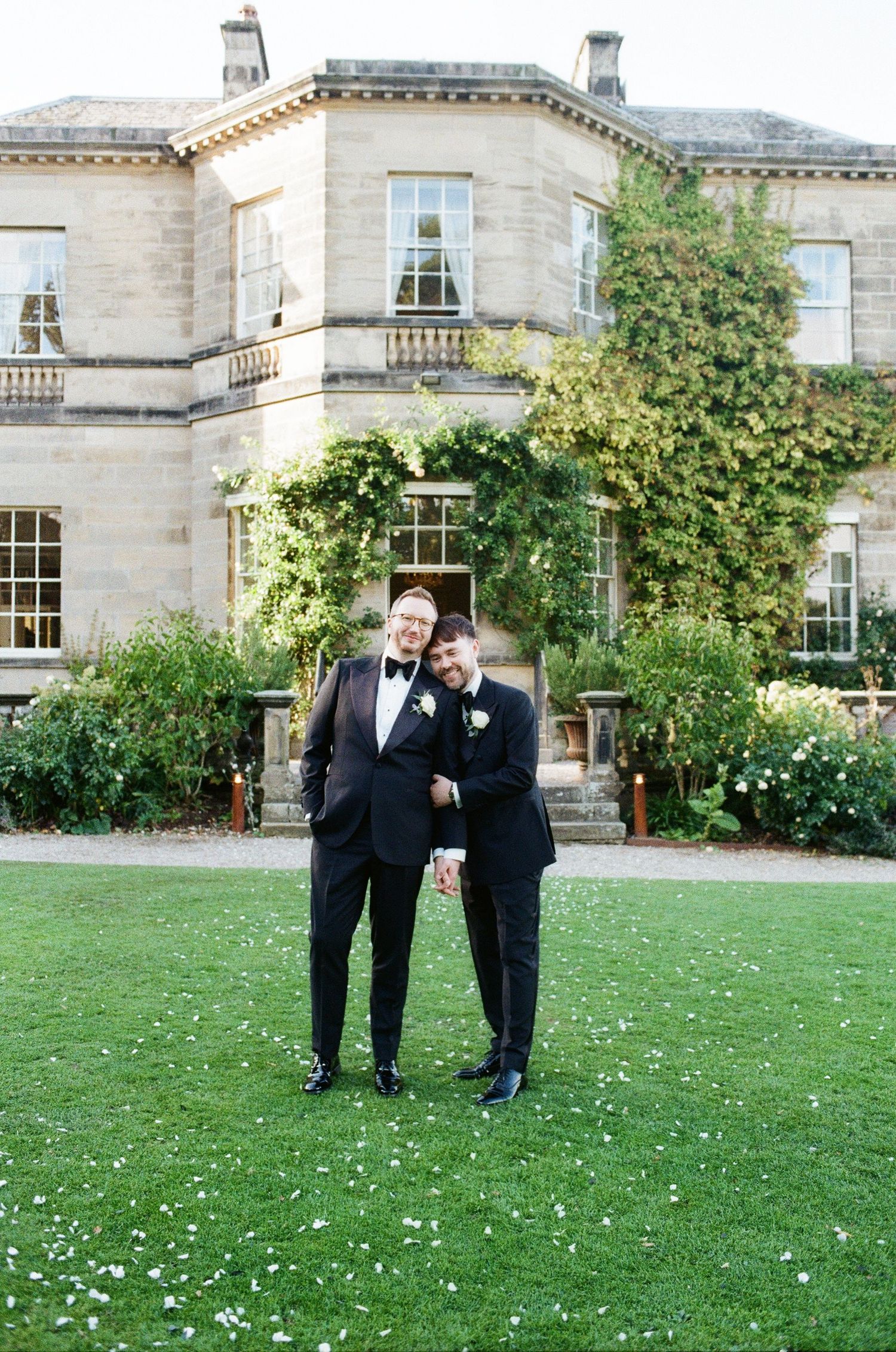 Two grooms in black tuxedos pose together on lawn in front of grand English country house wedding venue.