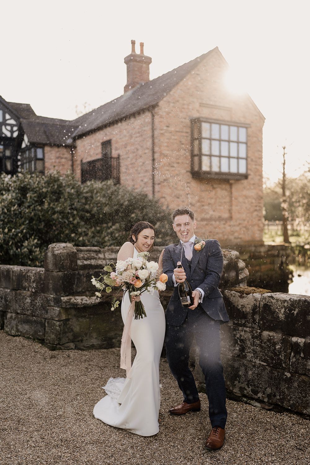 Bride and Groom standing laughing with a bottle of champagne spraying with Holford Hall behind.