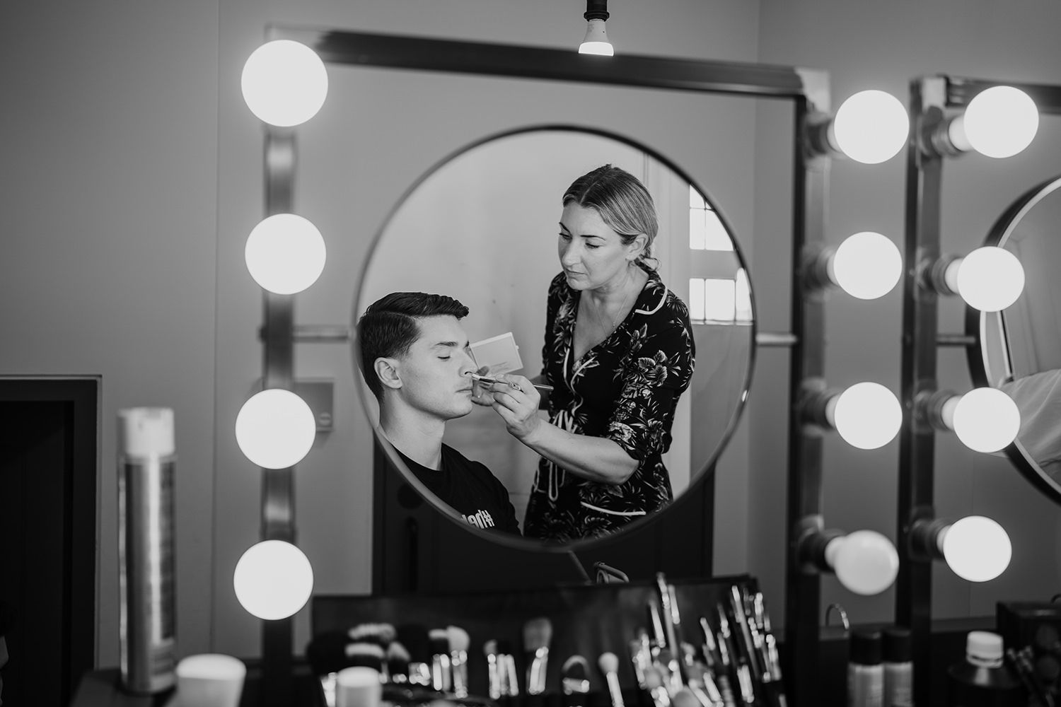Groom having his wedding makeup done in a reflection of a mirror surrounded by lights.