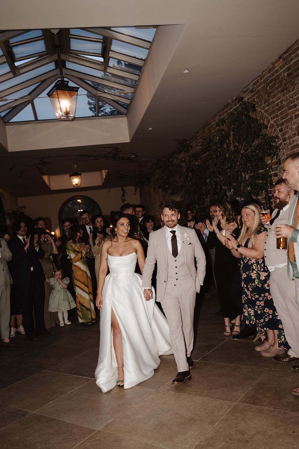 Newlyweds walk down a venue hallway with a glass ceiling and stone walls while wedding guests cheer and celebrate.