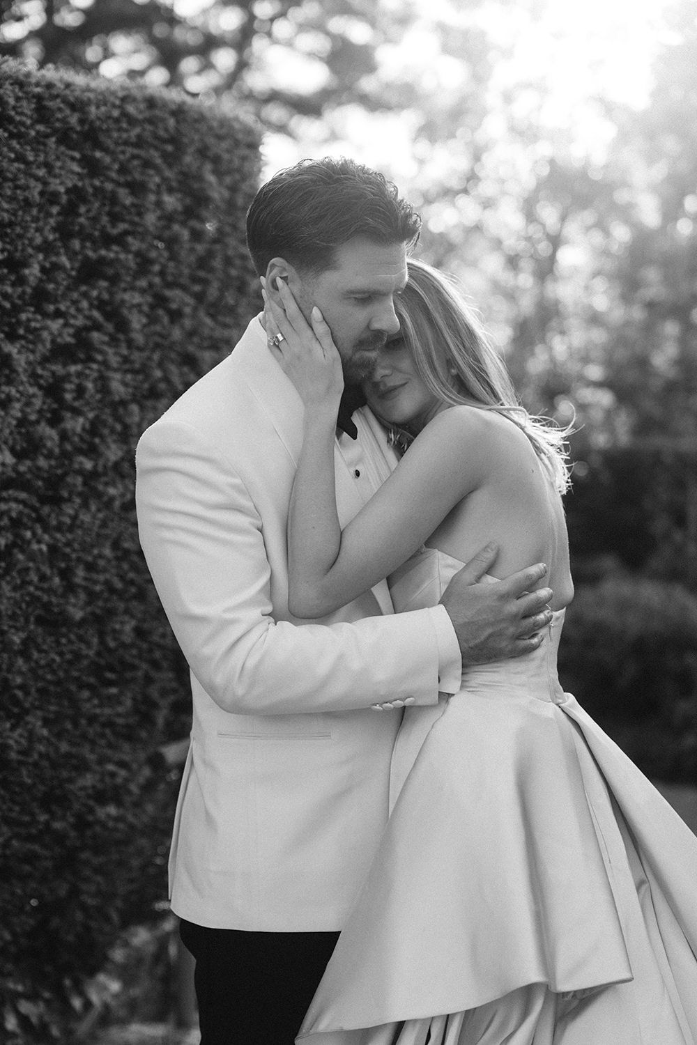 A tender moment between newlyweds sharing an intimate embrace in formal wedding attire against a garden backdrop.