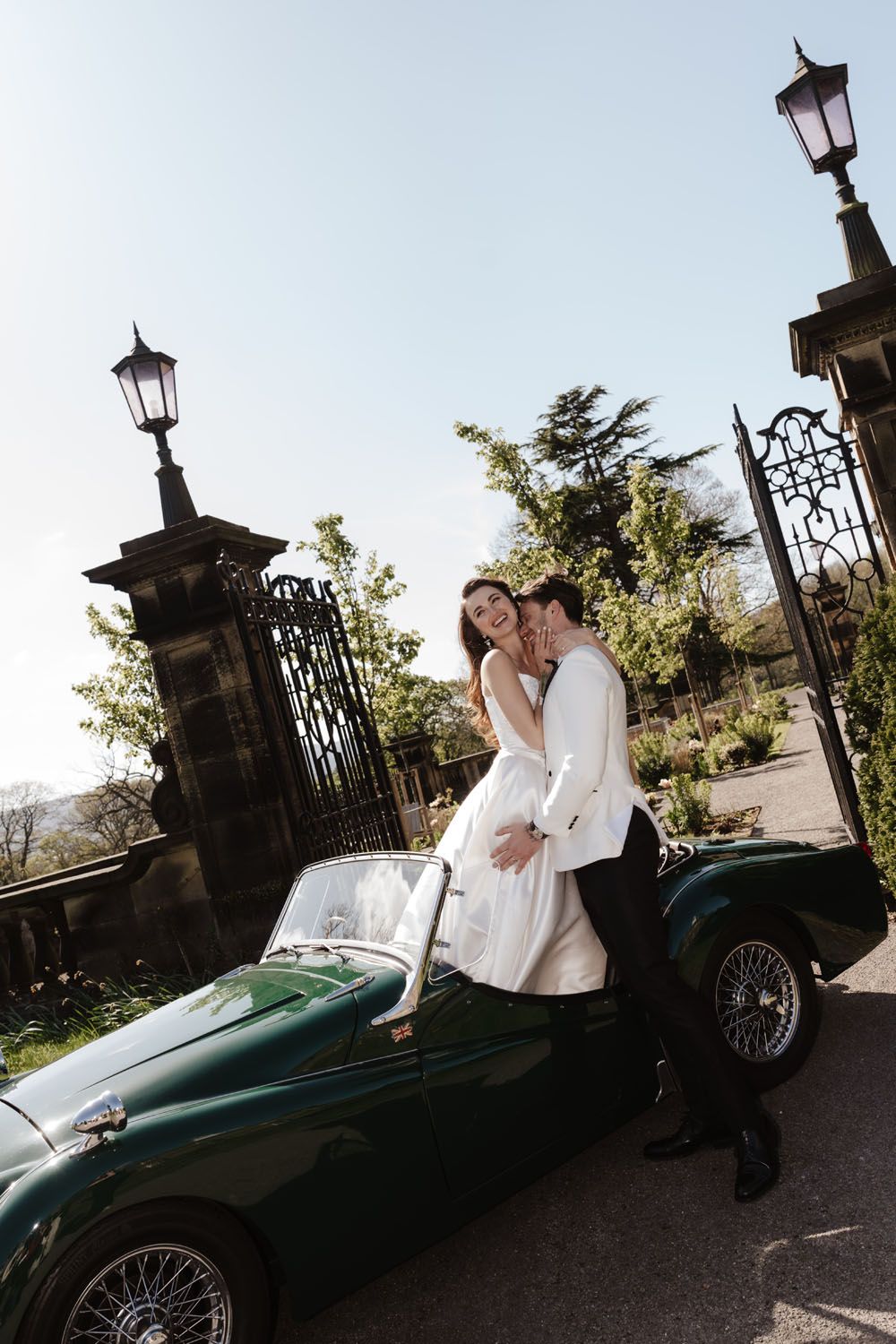 Bride and groom posing beside a vintage green convertible sports car at elegant iron gates.
