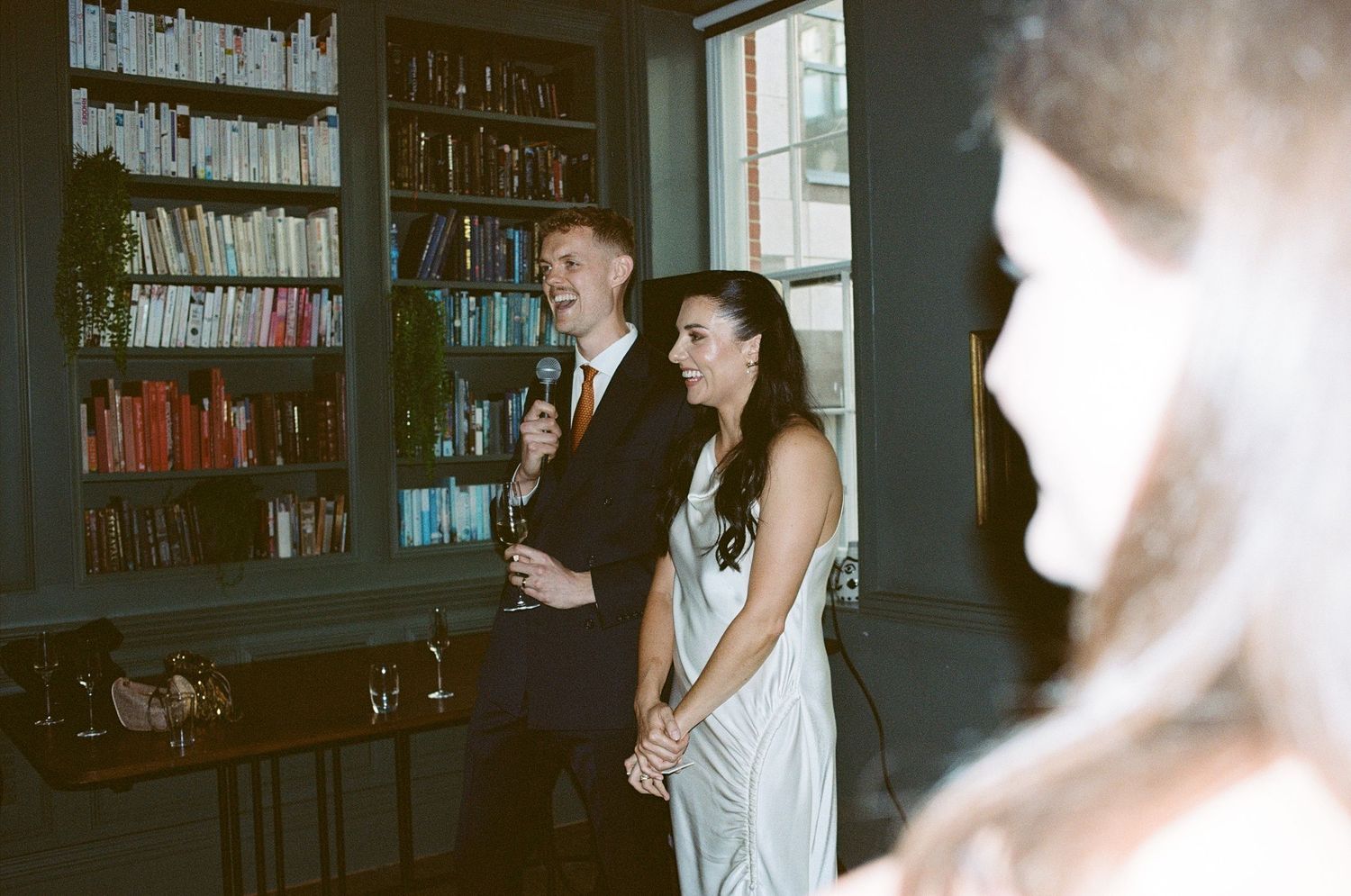 Couple stands together in a library venue during wedding reception, surrounded by bookshelves.