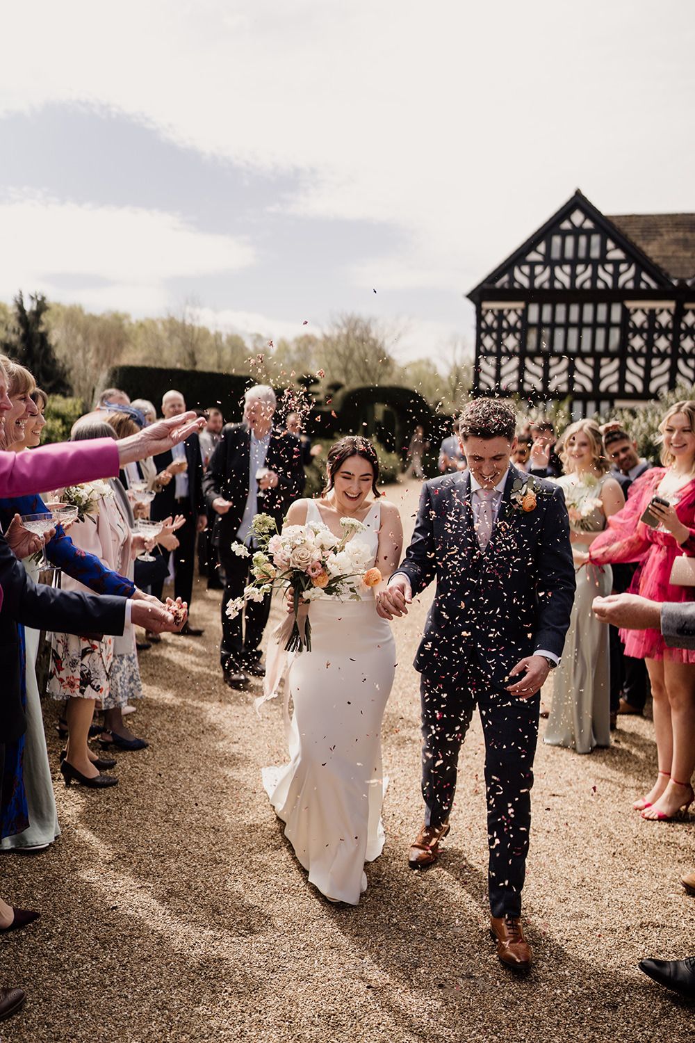 Black and white image of bride and groom walking amongst confetti with Holford Hall behind them as guests cheer.