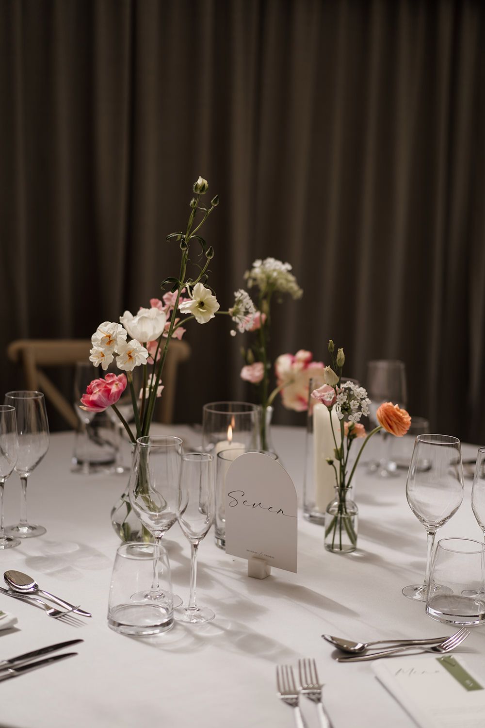Glass vases with colourful minimal wedding flowers, in the background there is a beige curtain backdrop.