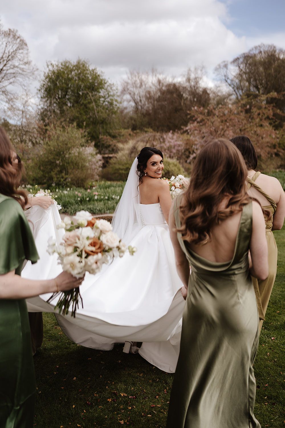Wedding party members in sage green dresses assist with a flowing white wedding gown outdoors on a spring day.