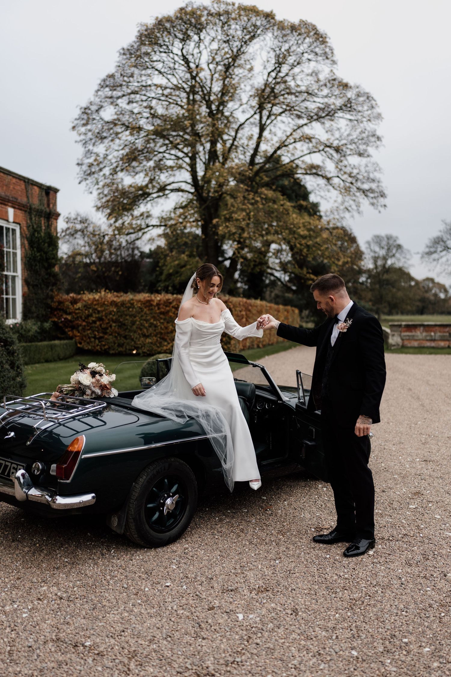 A classic vintage black sports car being used as a wedding vehicle on a gravel driveway near a historic building.
