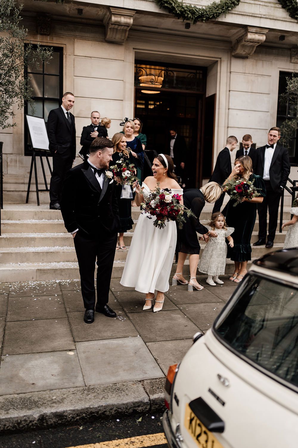 Wedding party gathered on stone steps outside venue with vintage car parked on street during intimate ceremony.