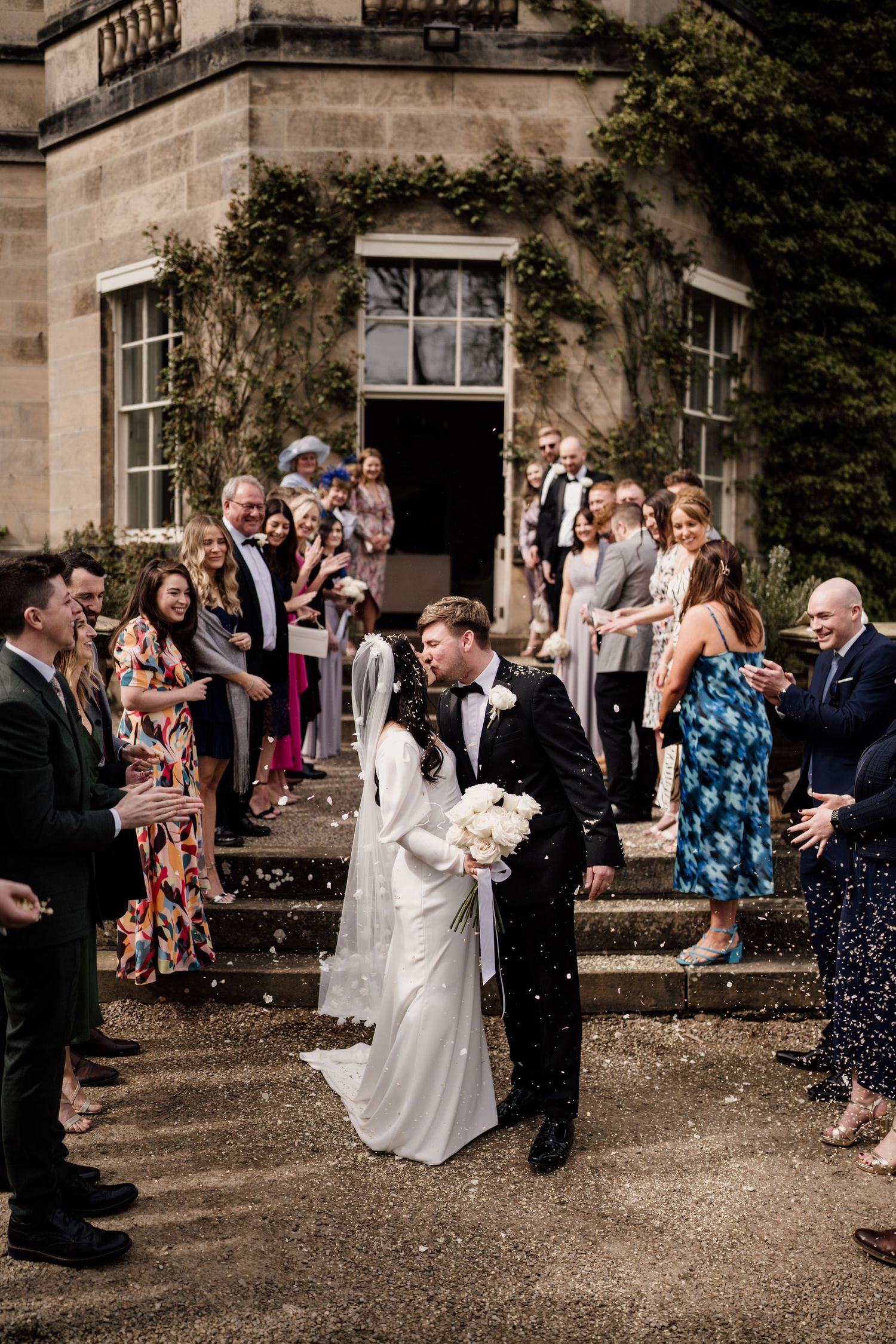 Bride and groom kissing as they have confetti thrown by friends and family.