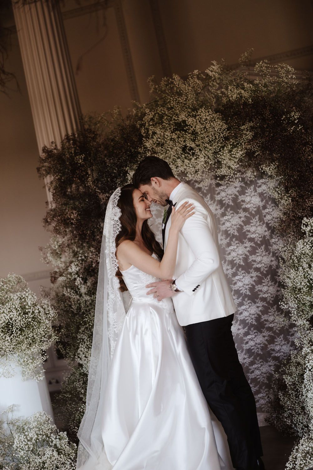 Bride and groom embrace tenderly before a lush baby's breath floral backdrop, bride wearing a white gown and veil.