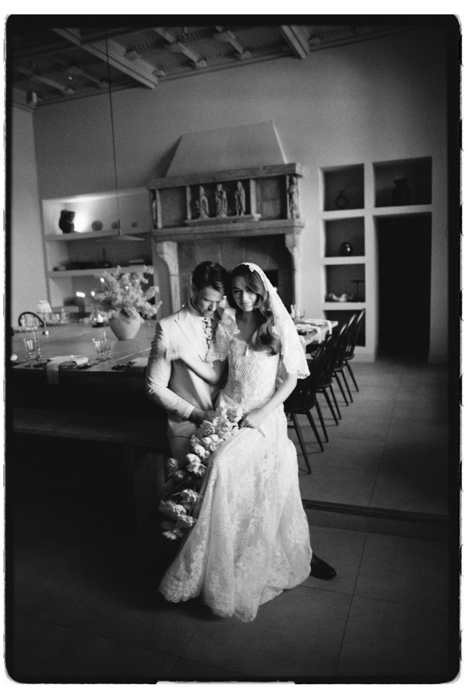 Black and white photo of a bride and groom in wedding attire holding flowers inside a rustic venue with fireplace.