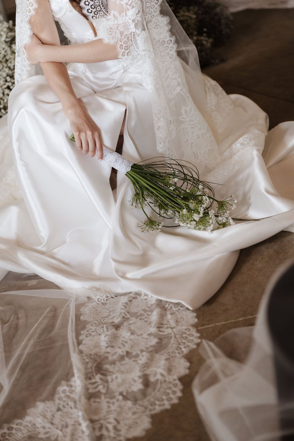 Bride in lace satin wedding gown seated with elegant white floral bouquet resting on dress train.