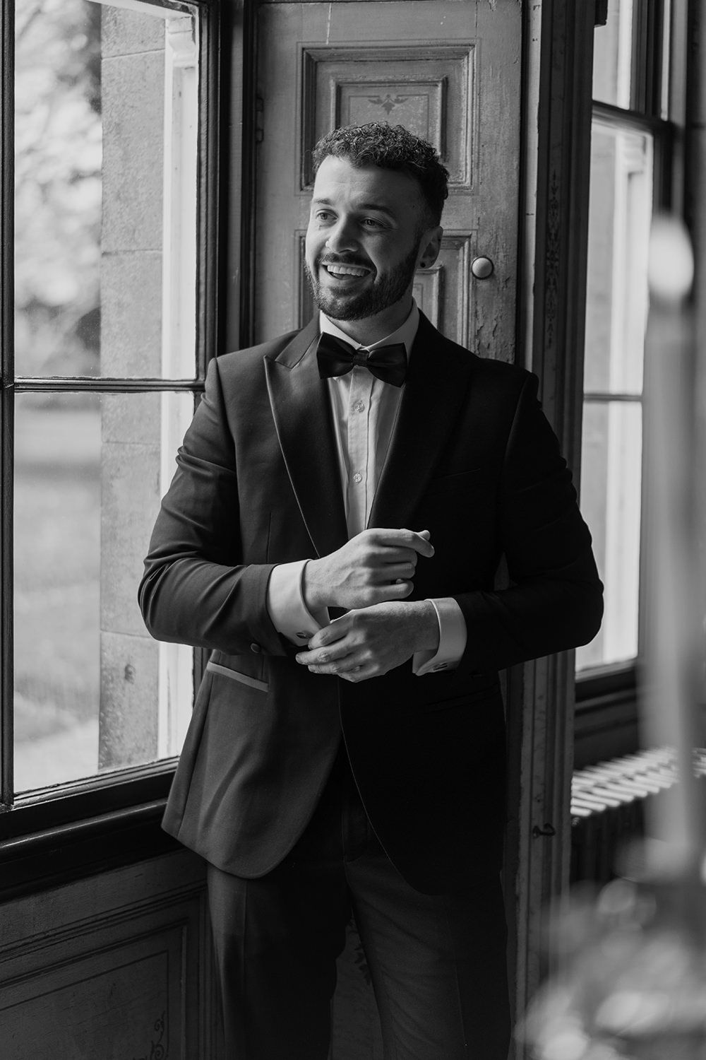 Groom adjusting his suit while smiling in the library at Rise Hall.