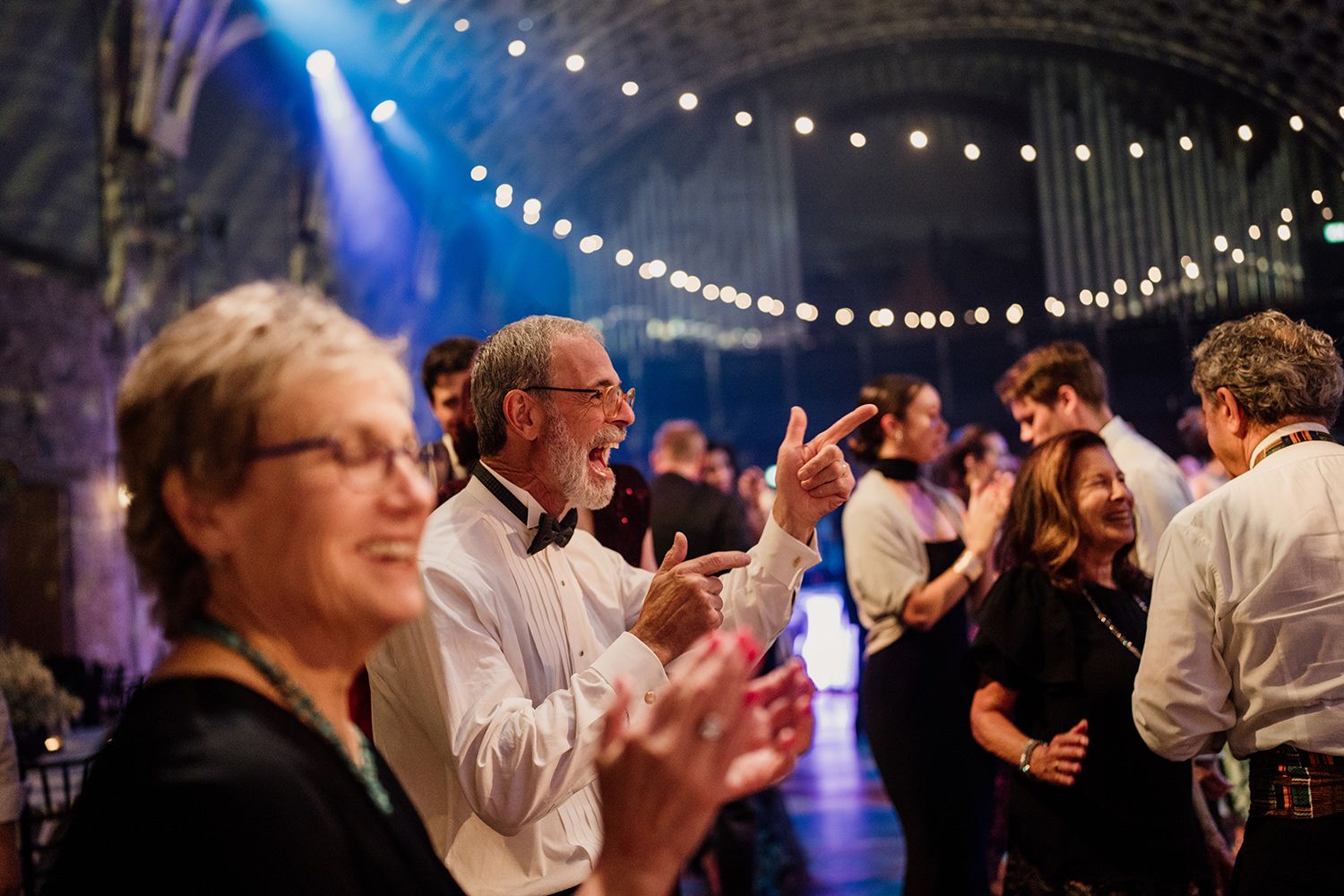 Wedding guests dancing, man is wearing a white shirt with a bow tie and pointing, disco lights are in the background.