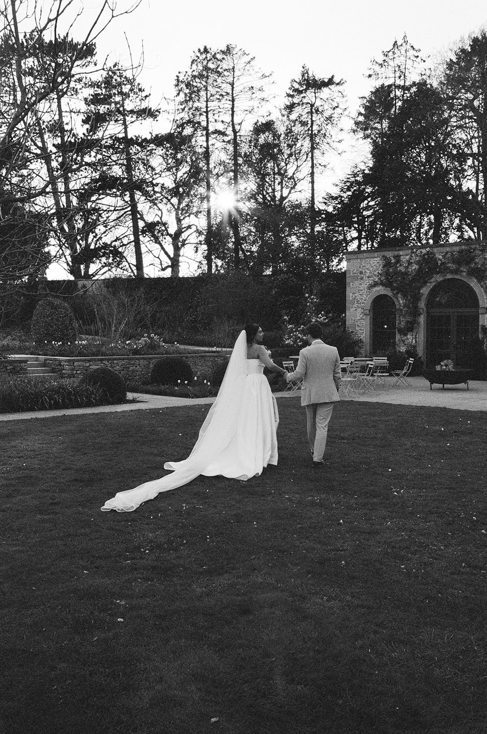 Black and white wedding photo showing a couple walking away across a lawn towards stone archways on a sunny day.