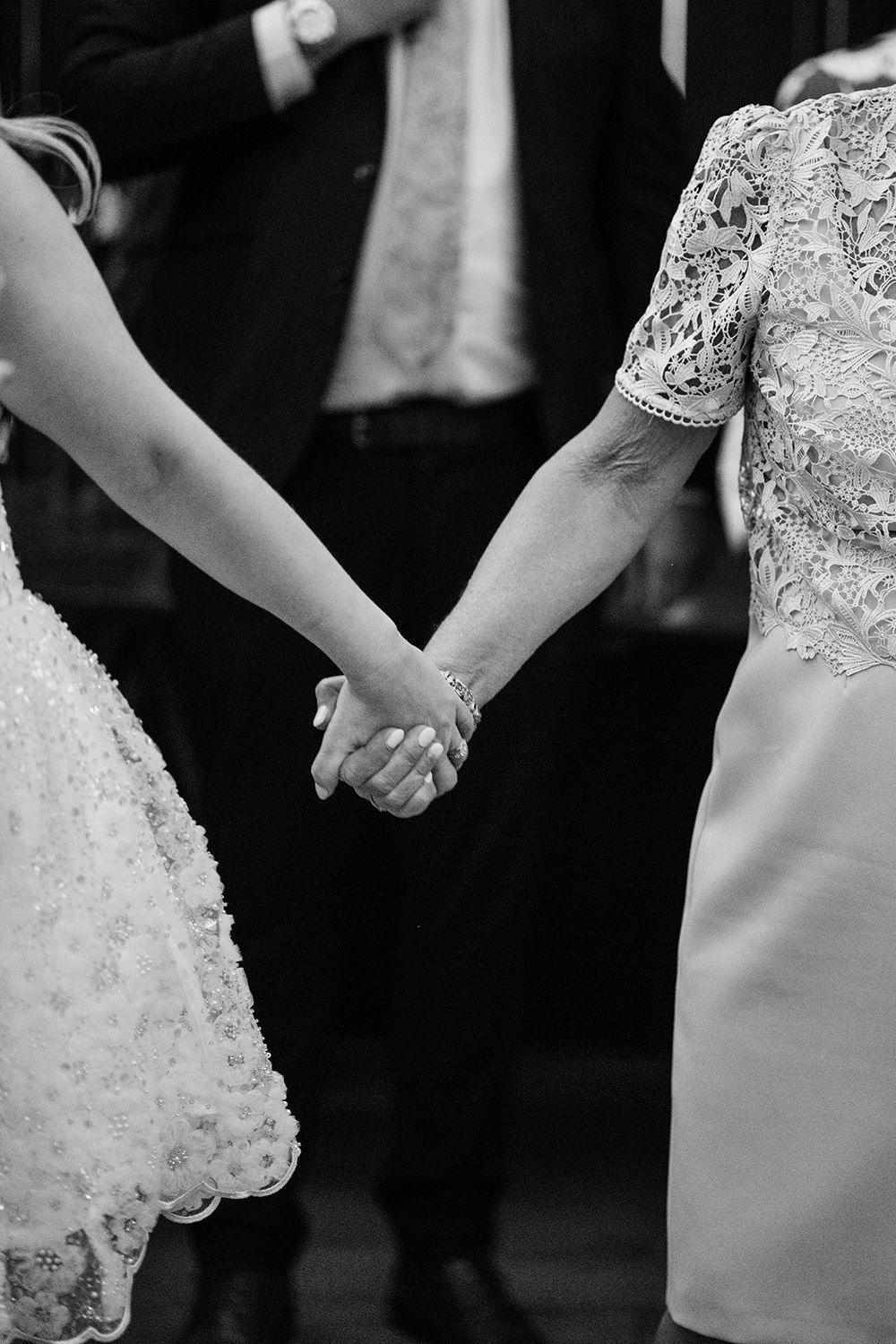 Bride and her mam holding hands, you can only see their arms and the background is black.