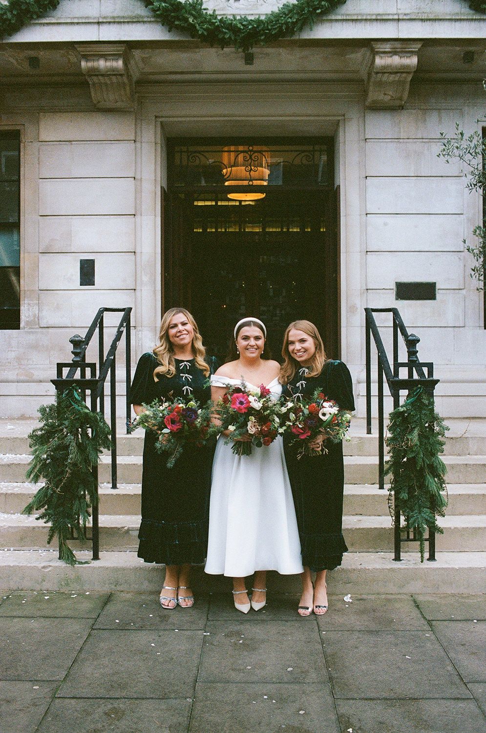 Bridal party stands at grand stone building entrance decorated with evergreen garlands and wreaths for winter wedding.