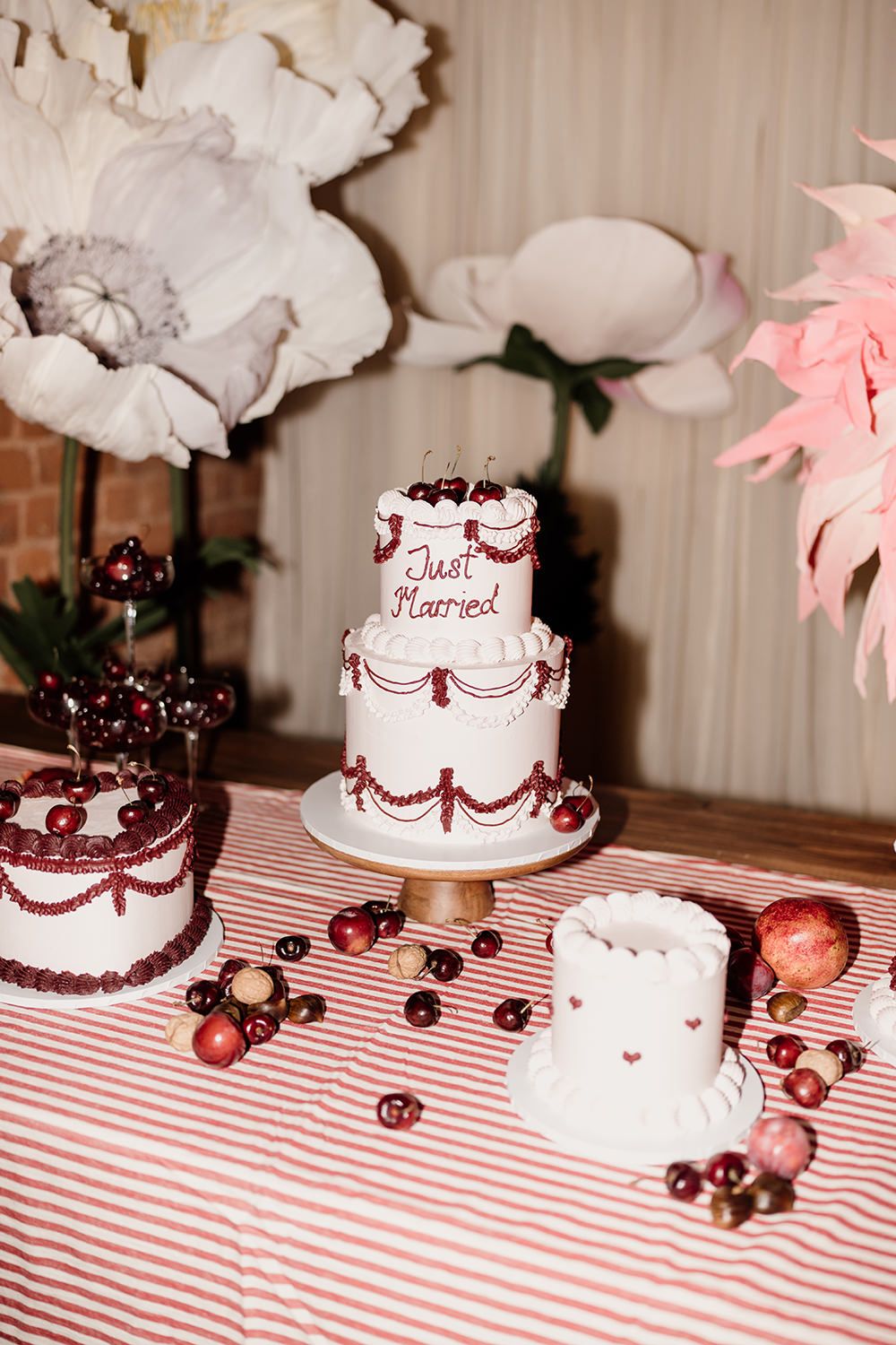 A white tiered wedding cake with burgundy decorative details sits on a striped tablecloth alongside scattered cherries.