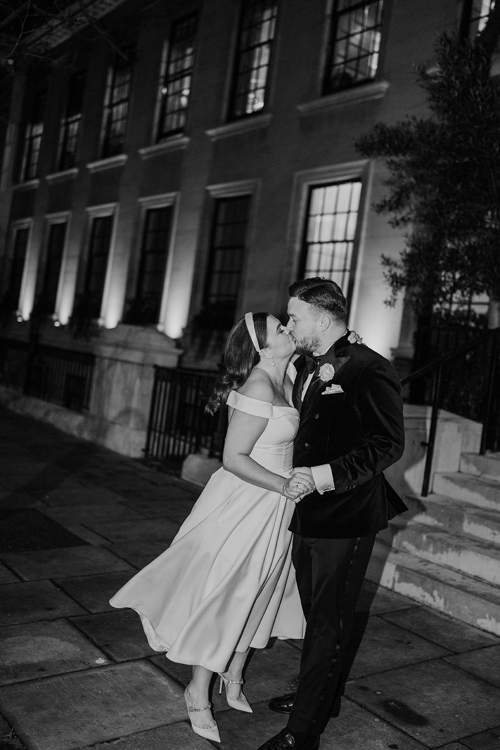A romantic black and white nighttime wedding photo showing a couple embracing on city steps beside a historic building.