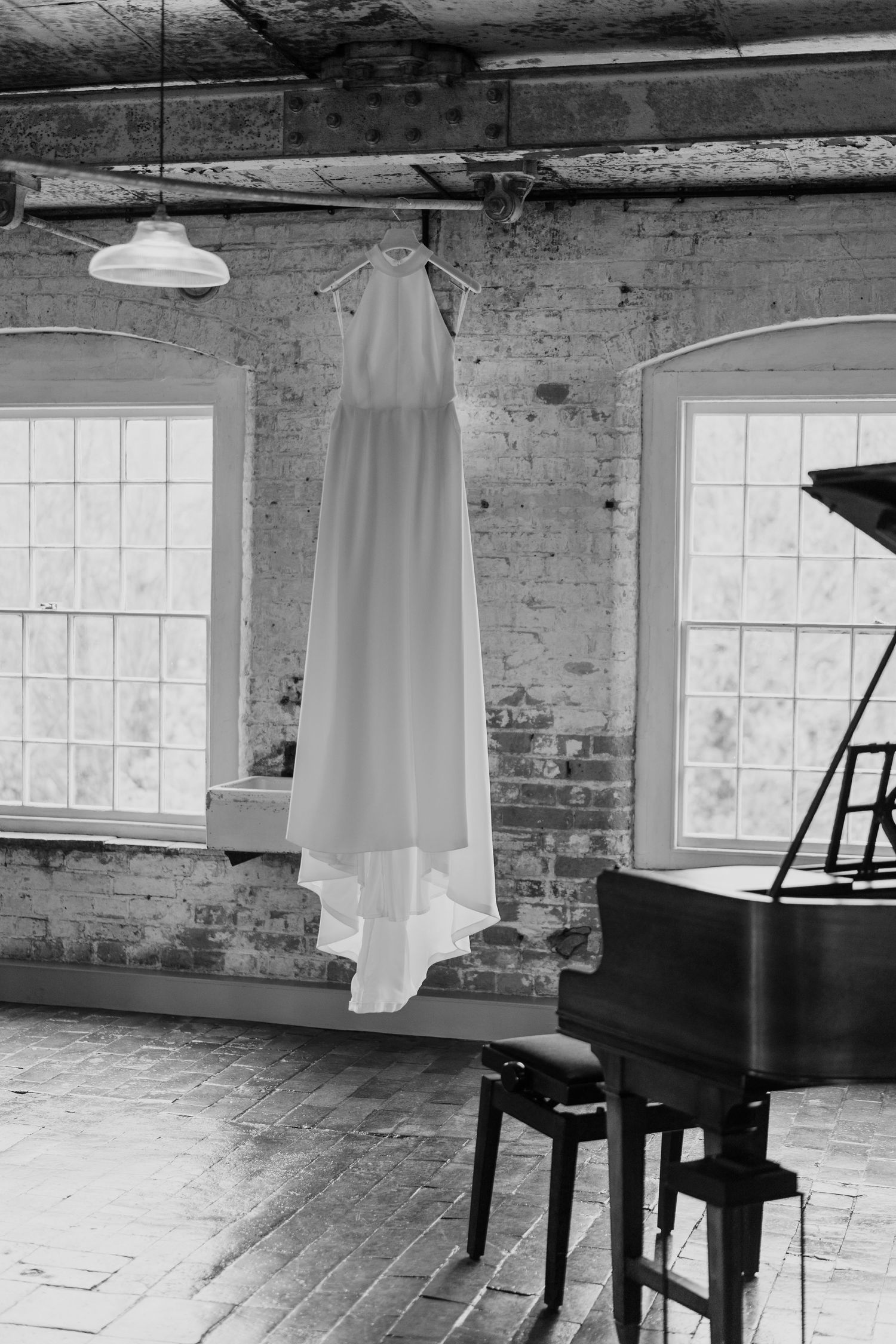 Wedding dress hanging in front of industrial windows with a piano in black and white.